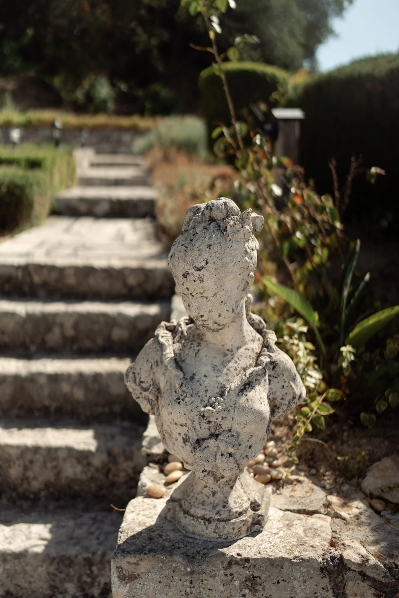 Weathered stone garden sculpture of a woman's bust with an elaborate necklace, placed at the base of stone steps in a landscaped garden.