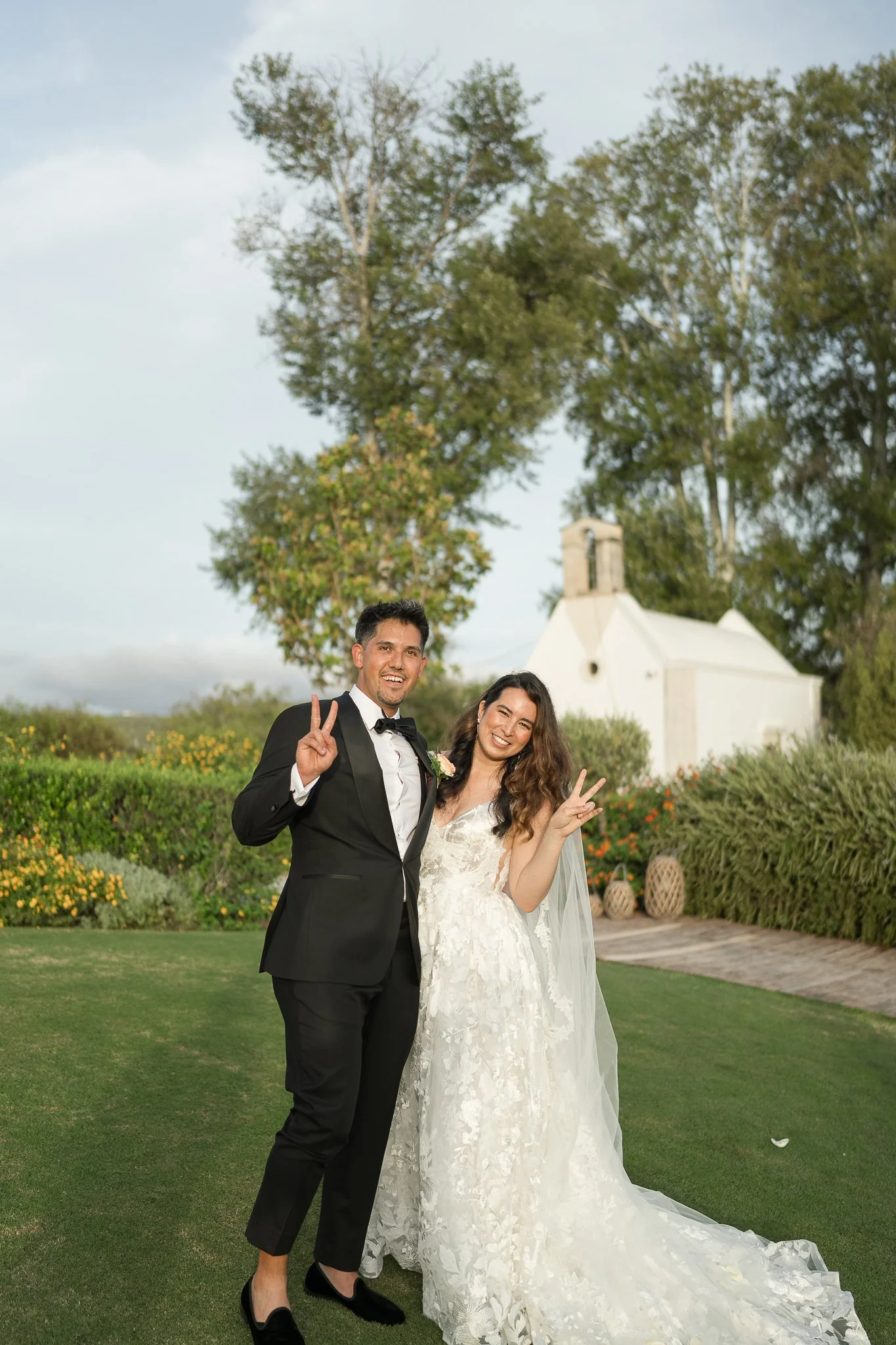 A newlywed couple in wedding attire smiling and making peace signs outside on a green lawn with trees and a white chapel in the background.