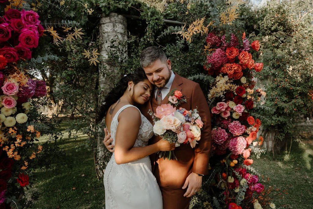 A bride and groom embracing under a floral wedding arch, with the bride holding a bouquet of pink and white flowers. The arch is decorated with vibrant pink, red, and orange flowers and lush green foliage.