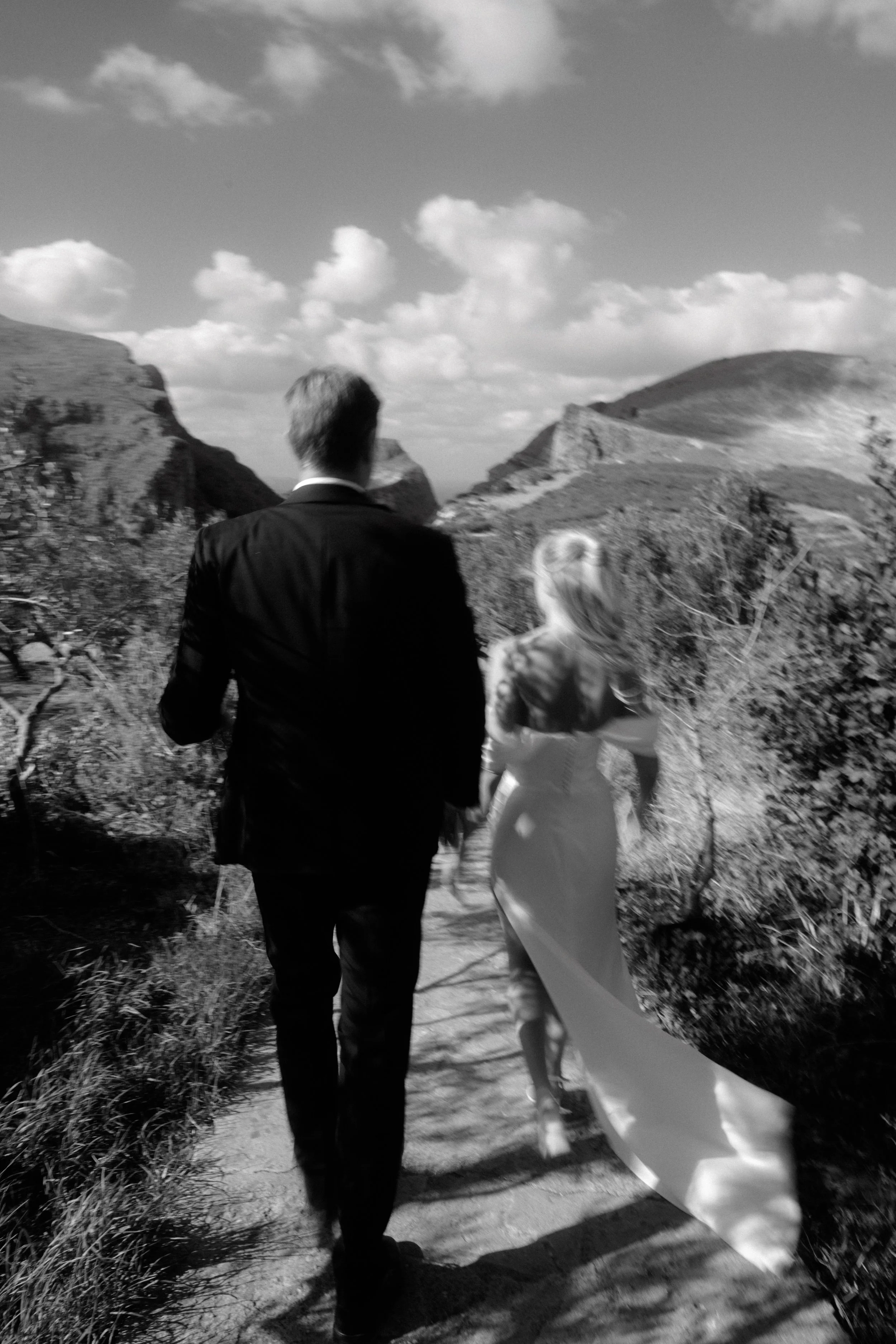 A black and white photo of a man in a suit and a woman in a wedding dress walking together on a path through rocky terrain with hills in the background.