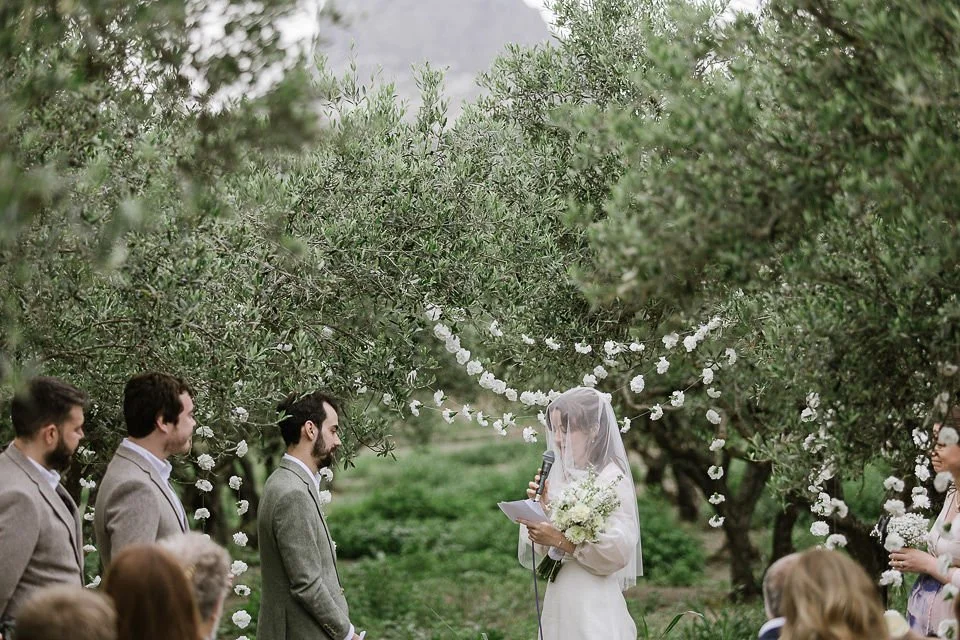 Bride in a white dress and veil reading vows during an outdoor wedding ceremony under an arch decorated with white flowers, surrounded by trees and guests.