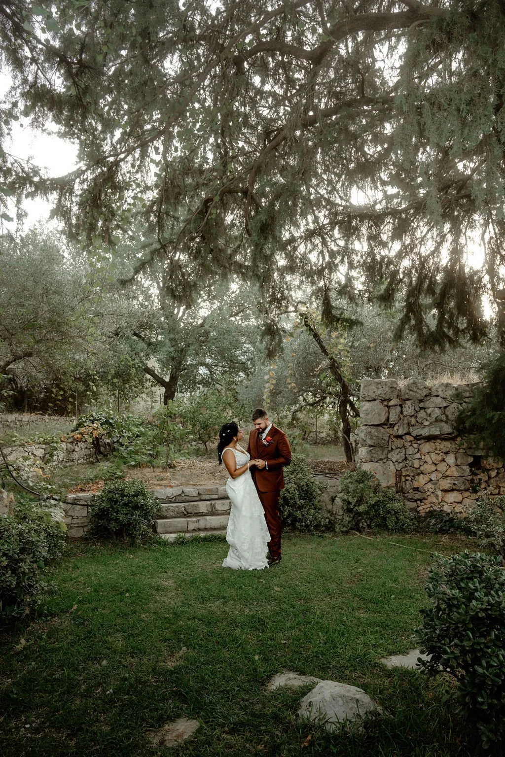A bride and groom standing together outdoors, holding hands, under large trees in a garden setting, during their wedding.