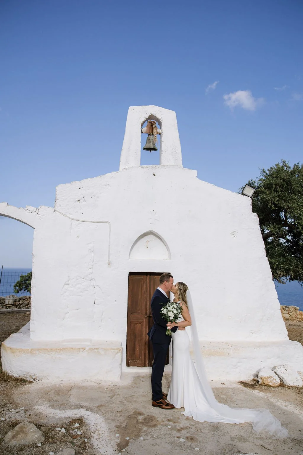A newlywed couple sharing a kiss in front of a white church with a bell and blue sky.