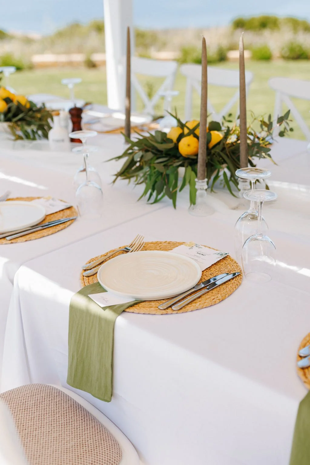 Elegant outdoor dinner table decorated with a white tablecloth, woven placemats, white plates, silverware, and upside-down wine glasses. A green cloth napkin is placed on each plate. Centerpiece with lemons, greenery, and tall candles is visible in t