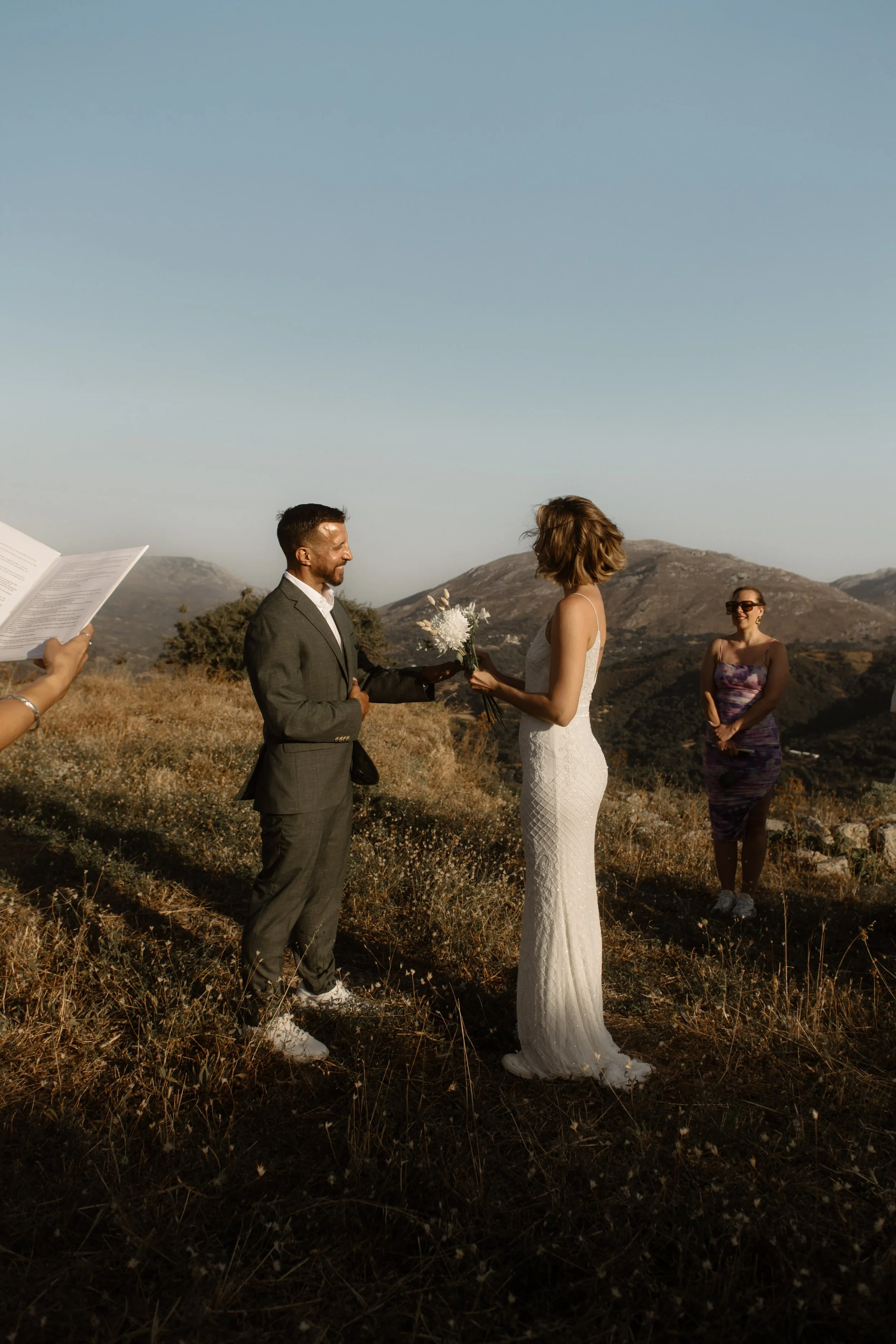 A couple getting married outdoors in a mountainous area, with a woman officiating and a woman in a floral dress watching, during late afternoon or early evening.