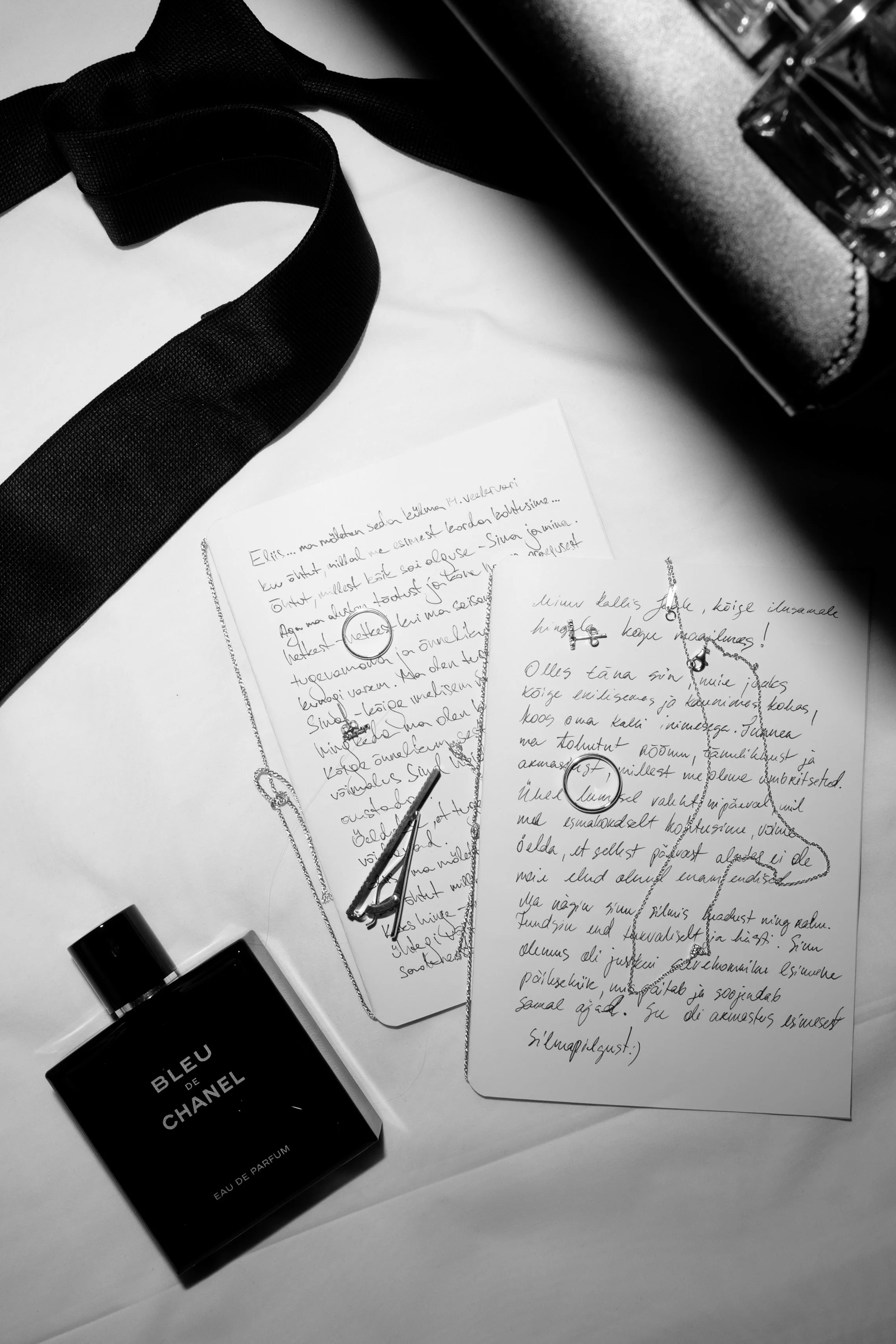 Black and white photo of two handwritten notes with necklaces placed on top, a bottle of Bleu de Chanel perfume, and a camera bag, all arranged on a desk or table.
