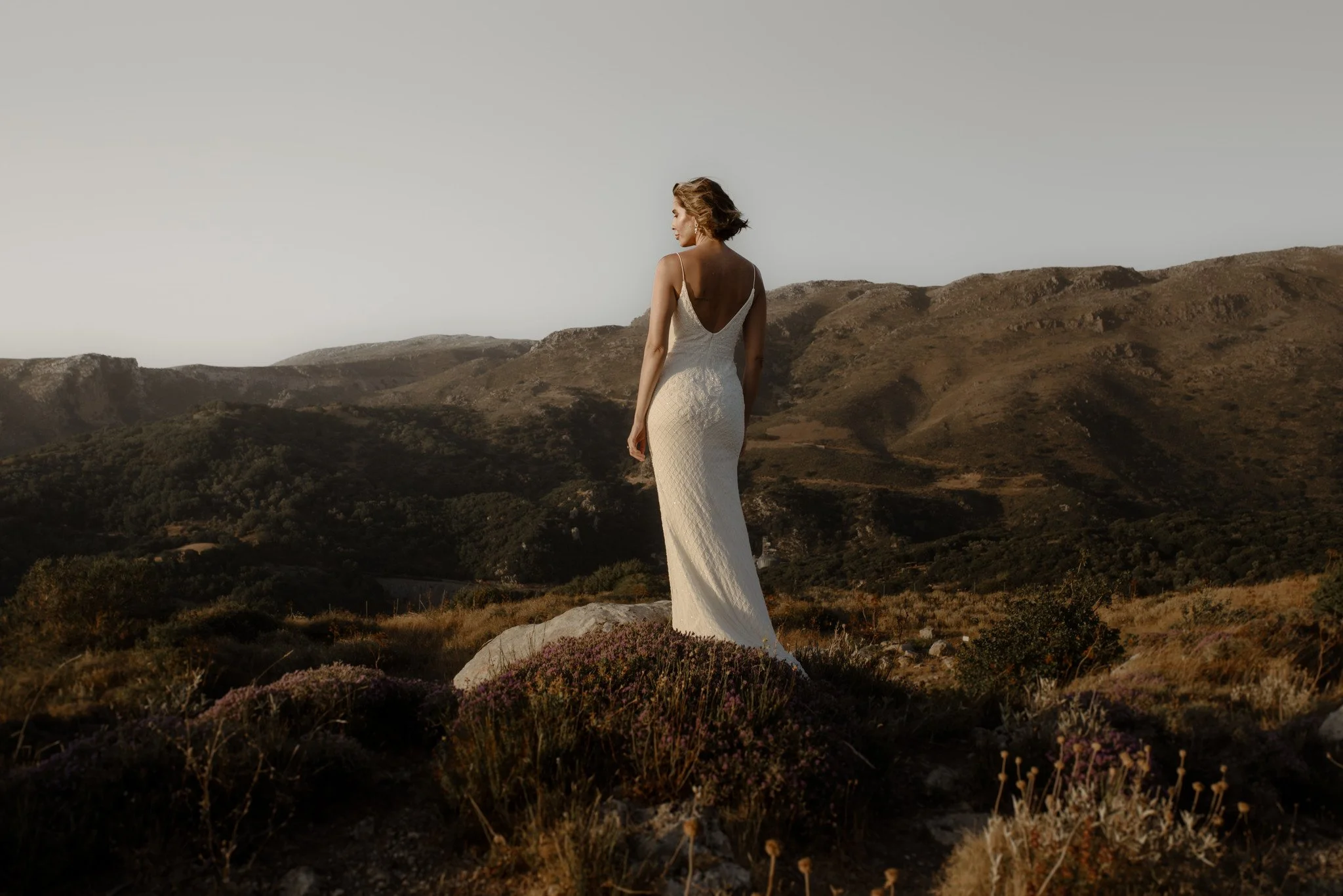 A woman in a white dress standing on rocky terrain in a mountainous landscape.