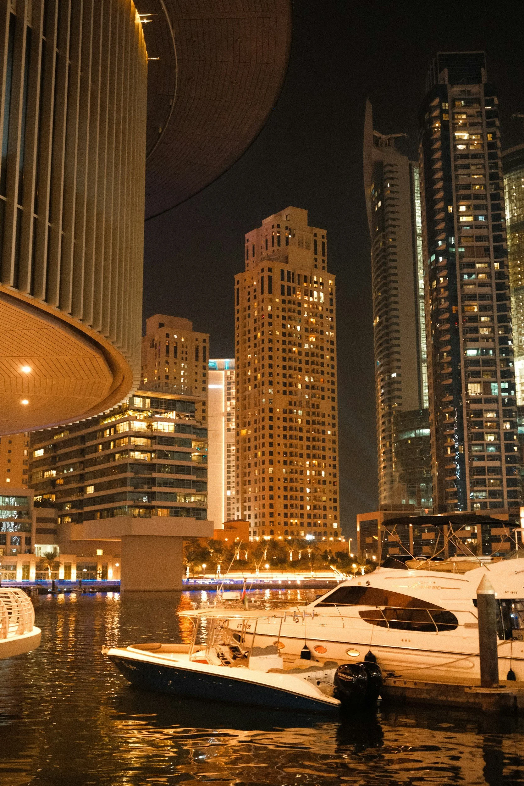 Night view of a city skyline with illuminated tall skyscrapers, reflecting on the water, with boats docked in the foreground.