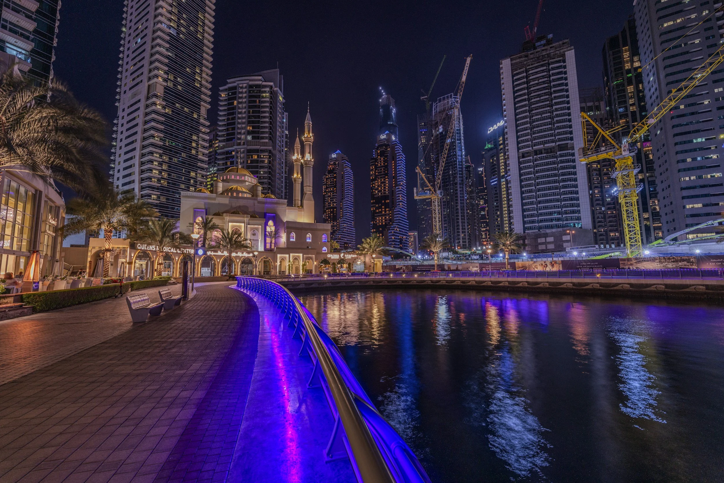 Nighttime view of Dubai Downtown with high-rise buildings, construction cranes, and a waterway illuminated with purple and blue lights, along a boardwalk with benches.