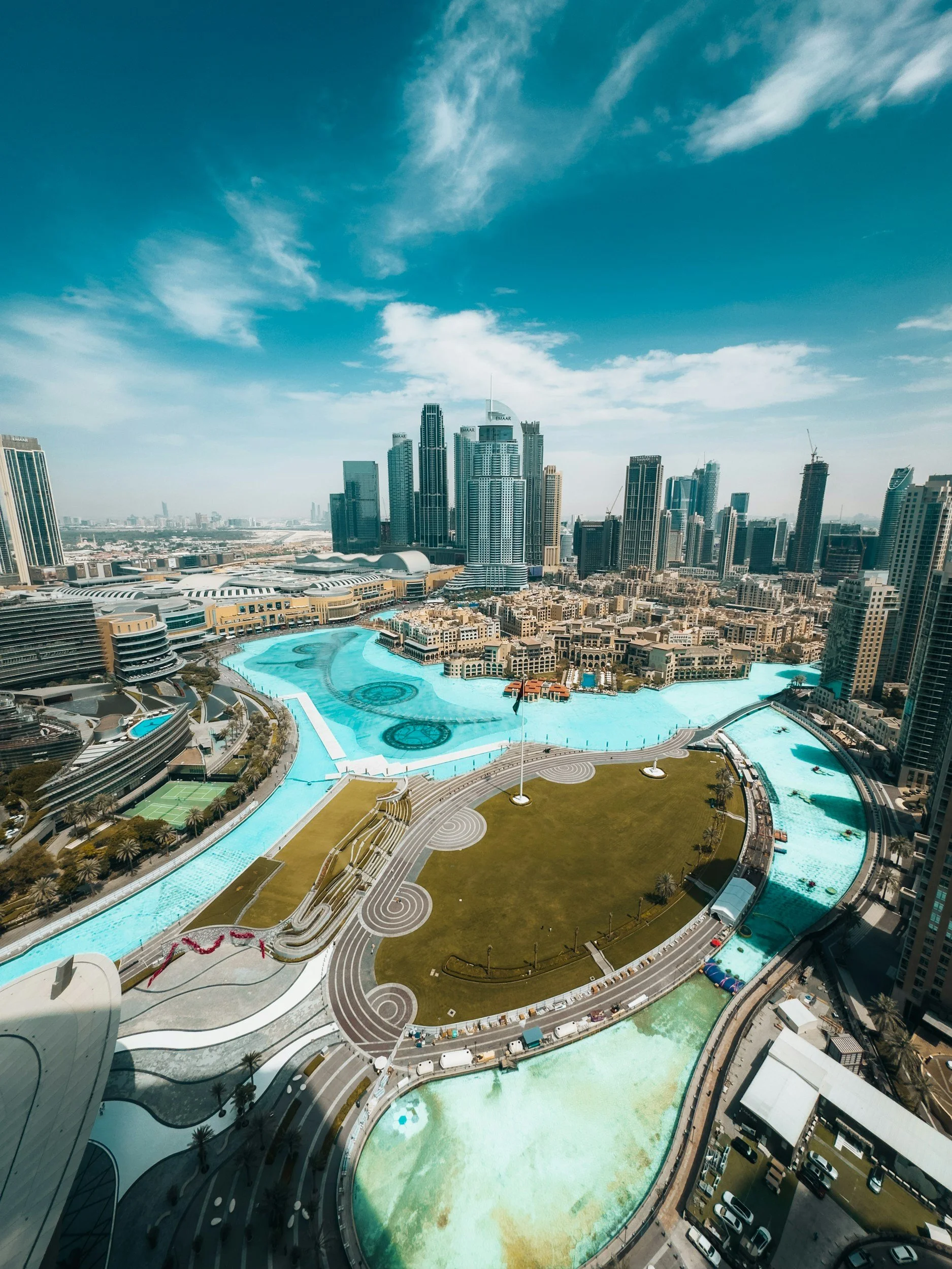 Aerial view of a modern cityscape with skyscrapers, a large pool or water feature, a green lawn, and surrounding buildings under a partly cloudy sky.