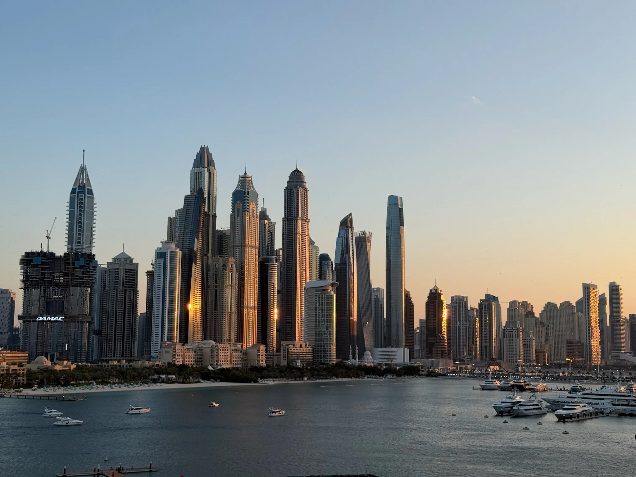 Skyline view of downtown skyscrapers along a waterfront, with boats docked in the harbor at sunset.