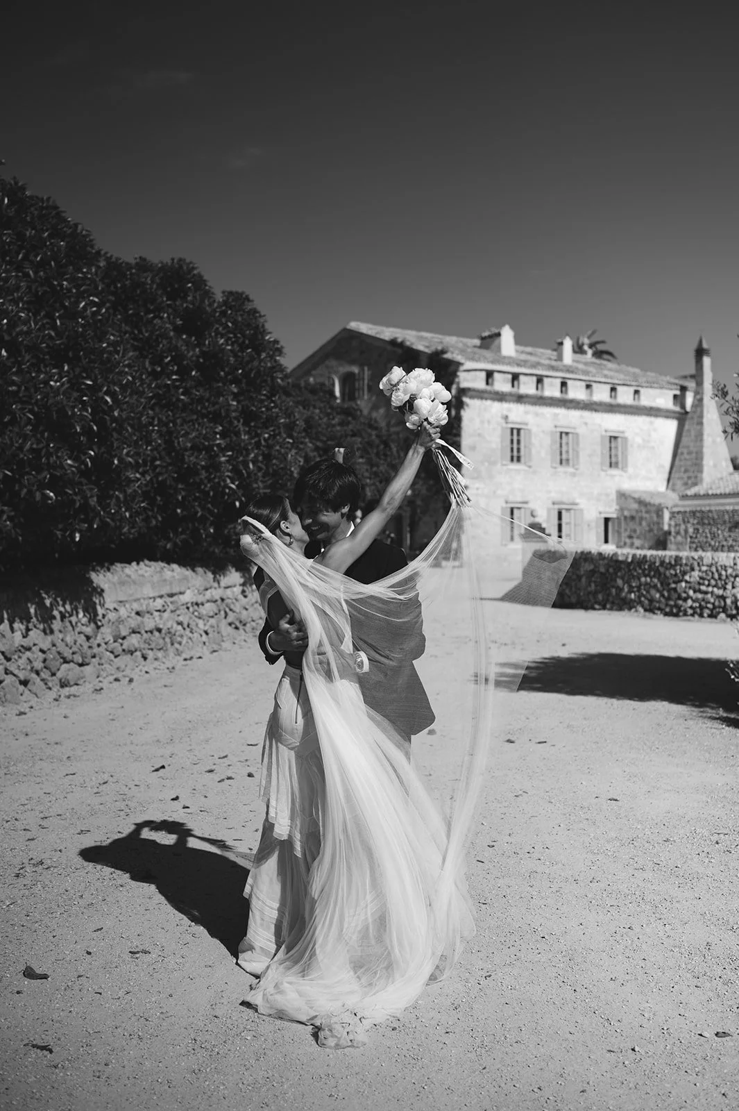 A black and white photo of a couple dancing outdoors, with one person lifting a bouquet of flowers. The scene includes a historic building and trees in the background.