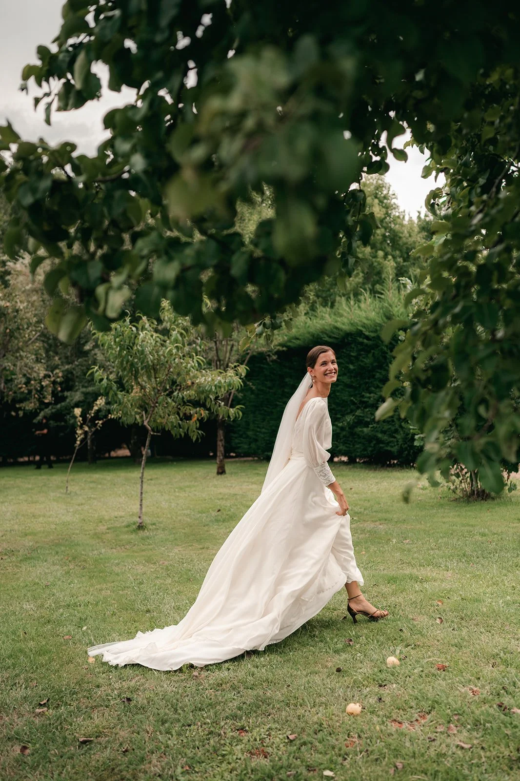 A bride in a white wedding dress smiling and walking in a lush green garden, framed by leafy branches.