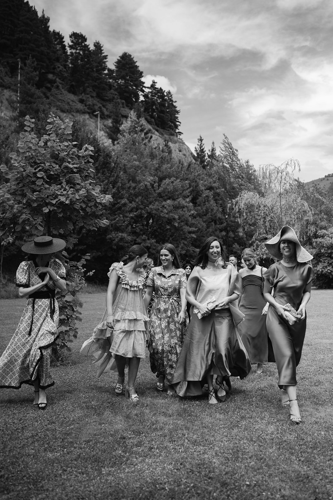 A group of women walking together outdoors in a scenic area with trees and hills, dressed in elegant dresses and wide-brimmed hats, smiling and enjoying themselves.
