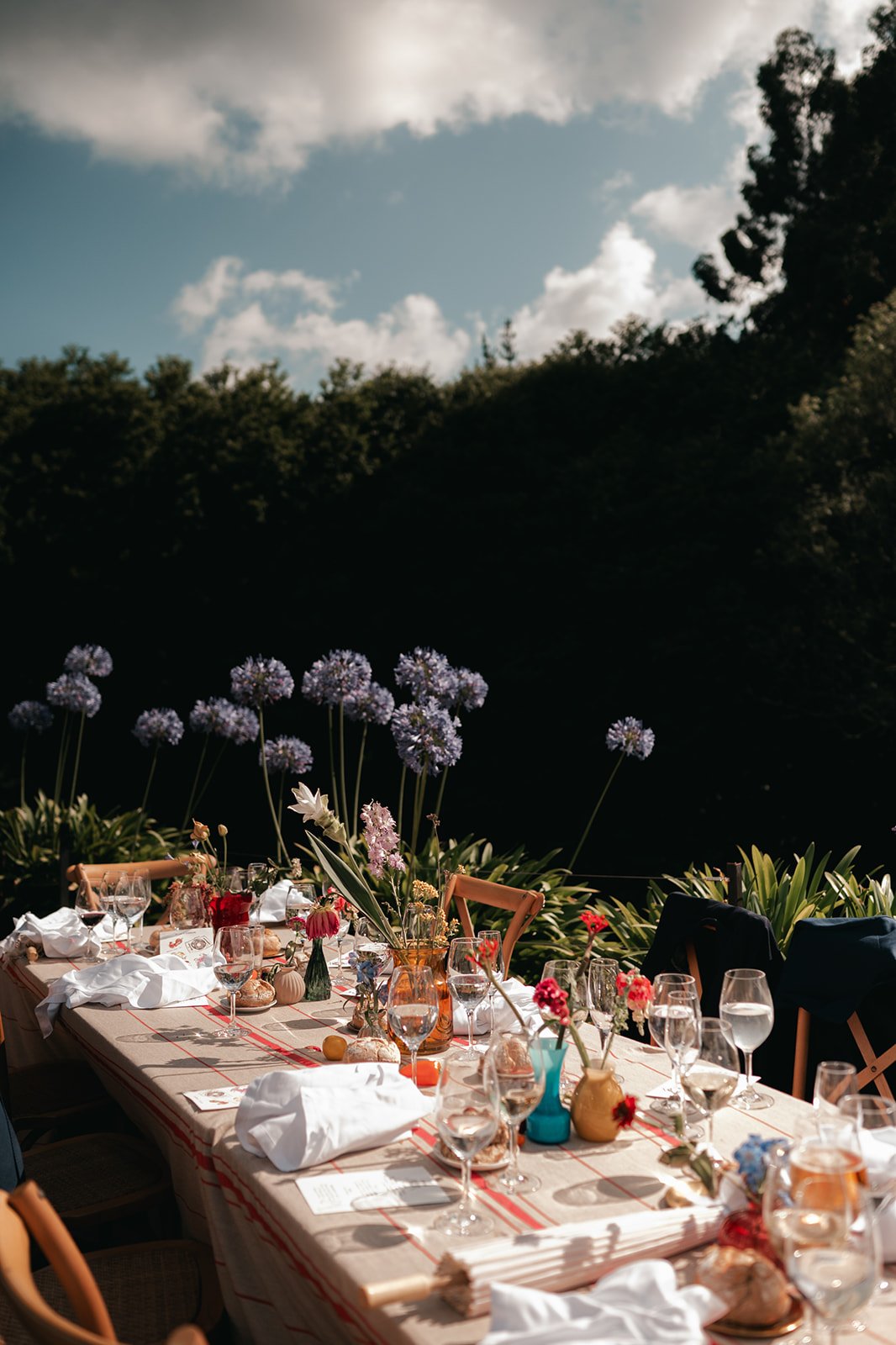 An outdoor dining table set with wine glasses, vases of colorful flowers, and tableware under a cloudy sky.