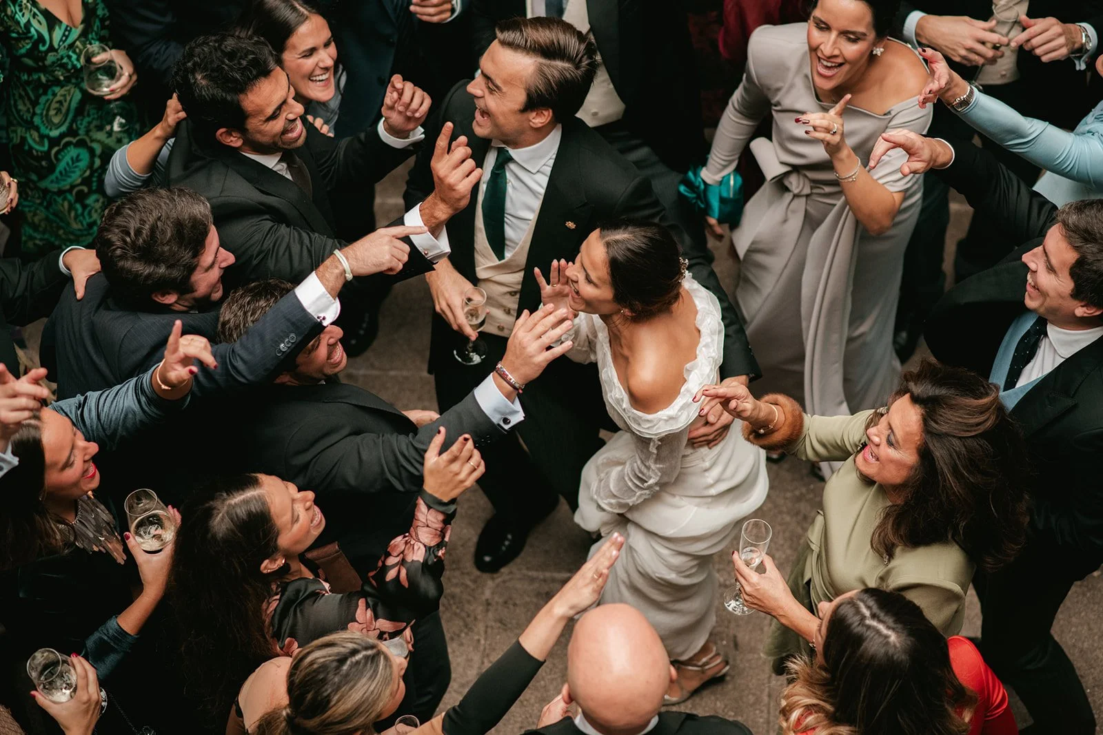 A group of people dressed in formal attire are dancing and celebrating at a wedding reception, with a bride in a white dress at the center.