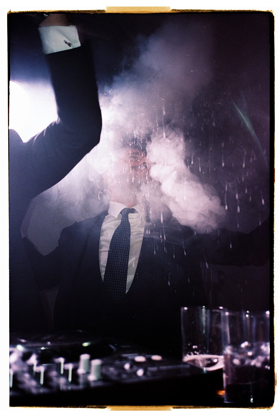 A man in a suit and tie is standing behind a glass window, with a cloud of powder or smoke in front of the glass, creating a foggy effect. A hand, likely of a DJ or performer, is holding a white object close to the glass. There are drinks and a DJ controller on the table in front of him.