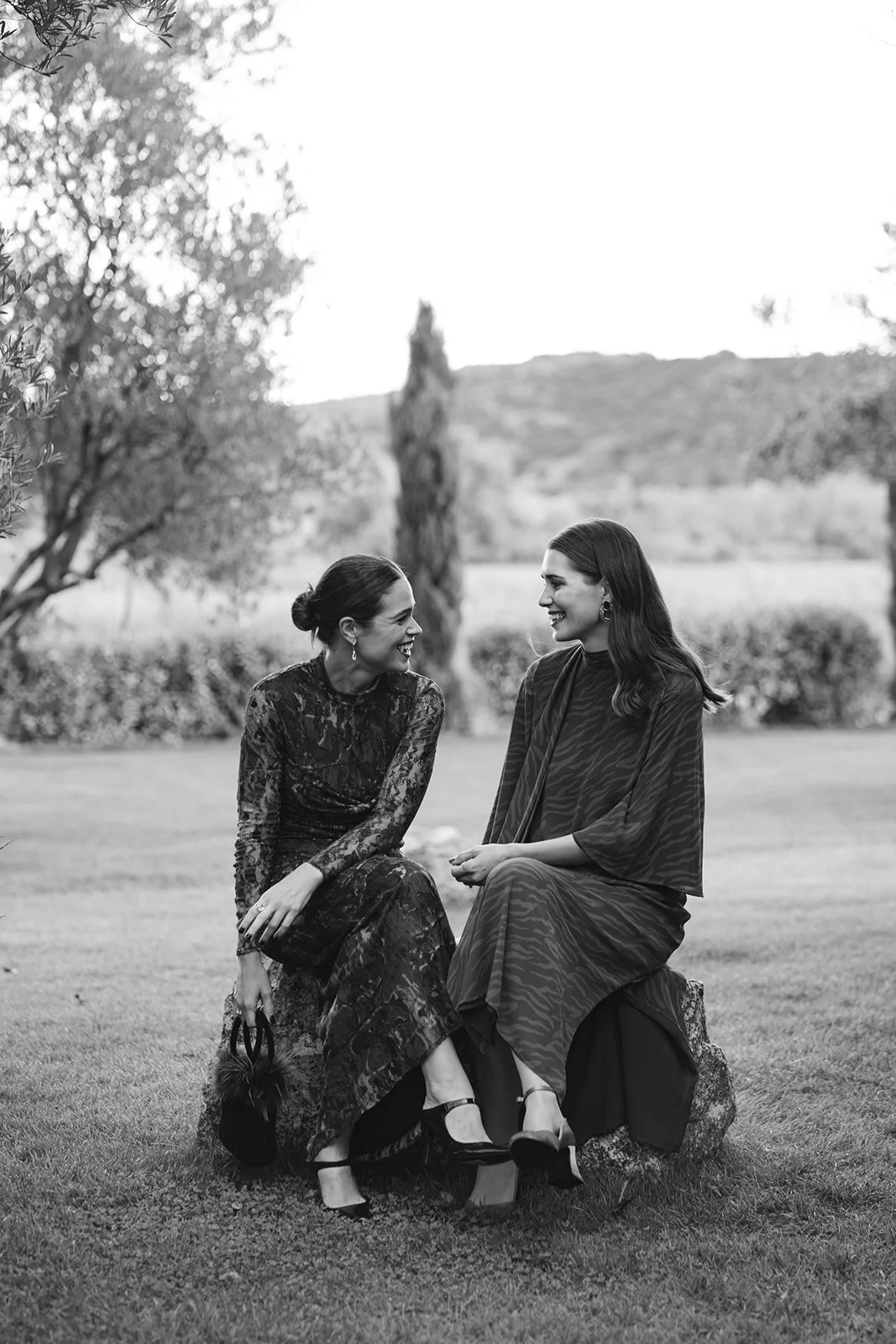 Two women sitting on a rock outdoors, smiling and engaging in conversation, with trees and hills in the background.