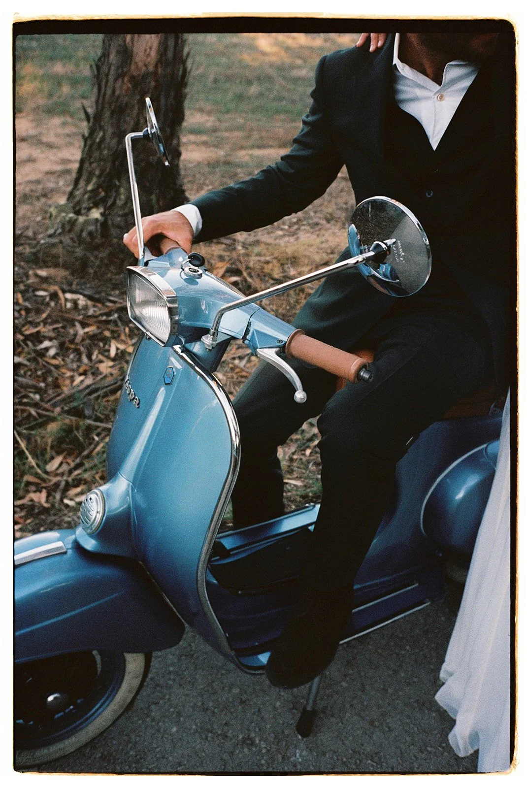 Man in a black suit riding a vintage blue scooter with brown leather handles on a dirt path in a wooded area.