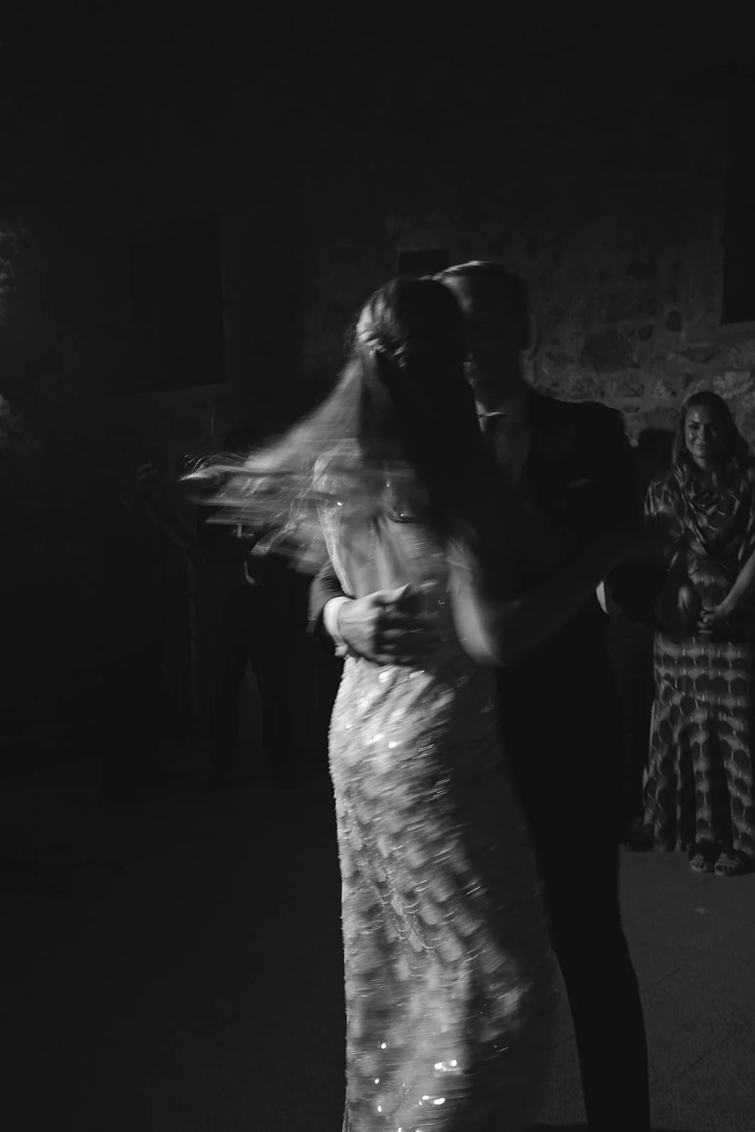 A couple dancing closely at a dimly lit event, with other guests watching and smiling in the background.