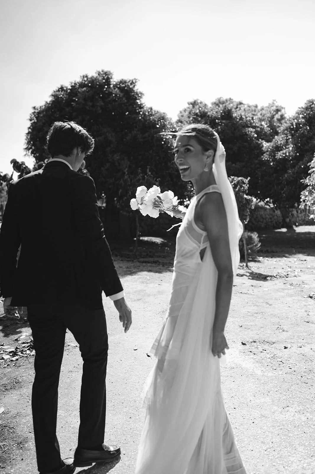 A bride in a white wedding dress and a groom in a black suit smile and laugh outdoors. The bride holds a bouquet of flowers, and both are standing on a dirt path with trees in the background.
