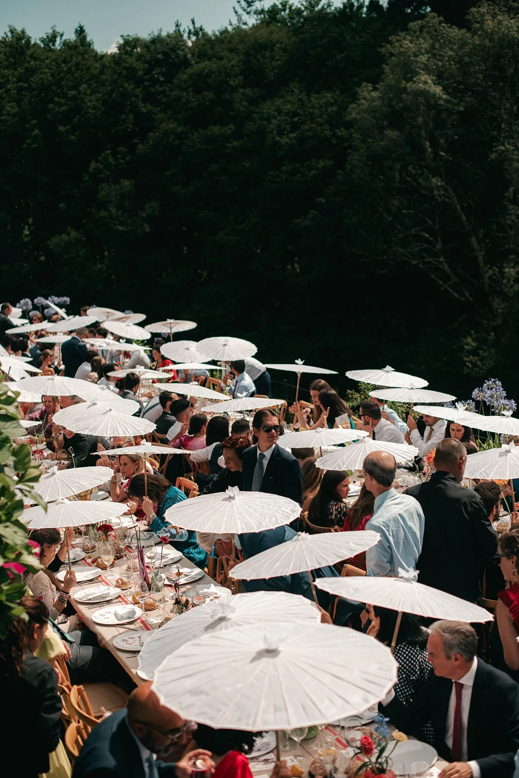 Outdoor wedding or event reception with long banquet tables, white parasols, and guests seated under a forested area.