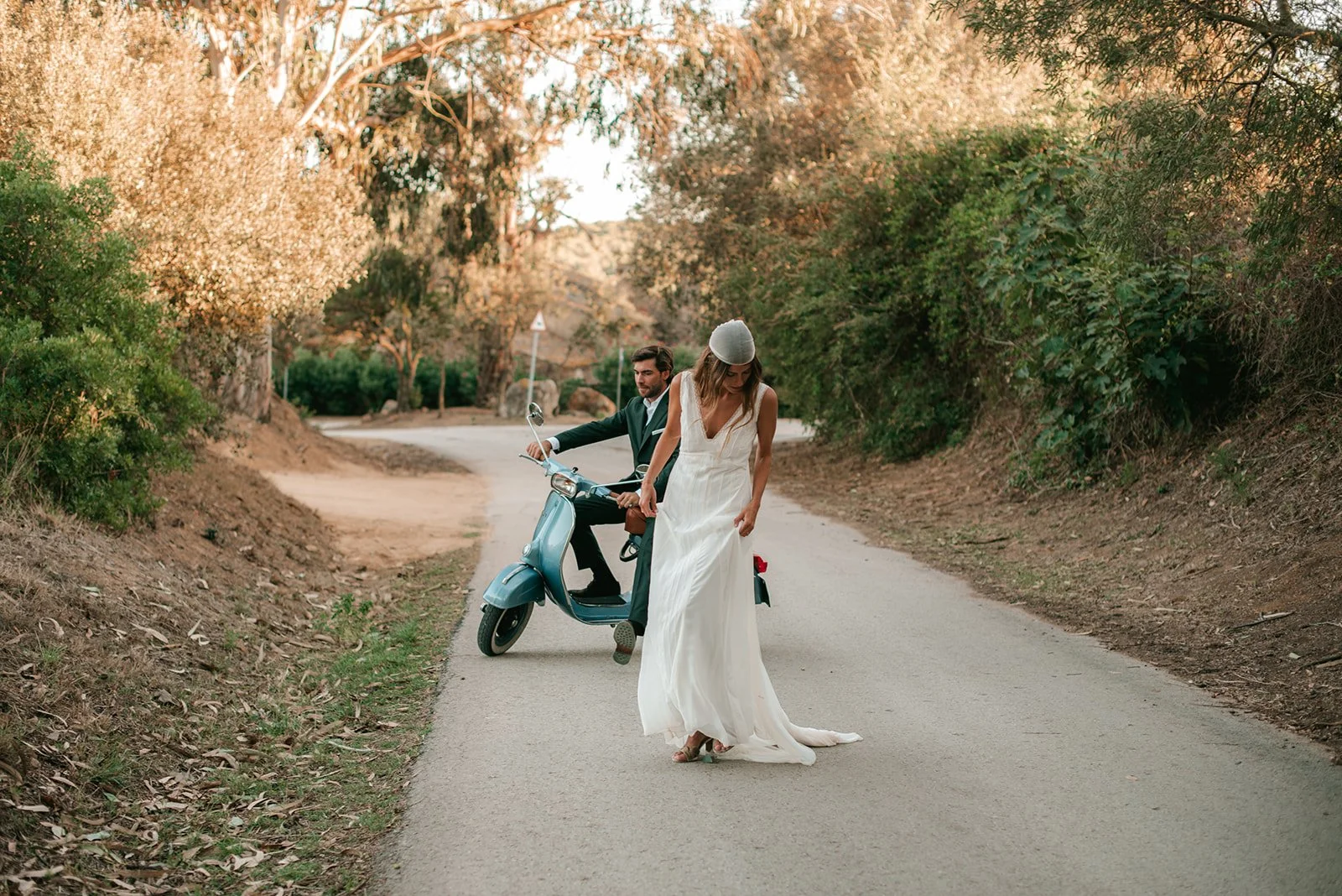A woman in a white dress and a gray hat standing on a deserted country road while a man in a suit rides a blue scooter behind her; surrounded by trees with autumn-colored leaves.