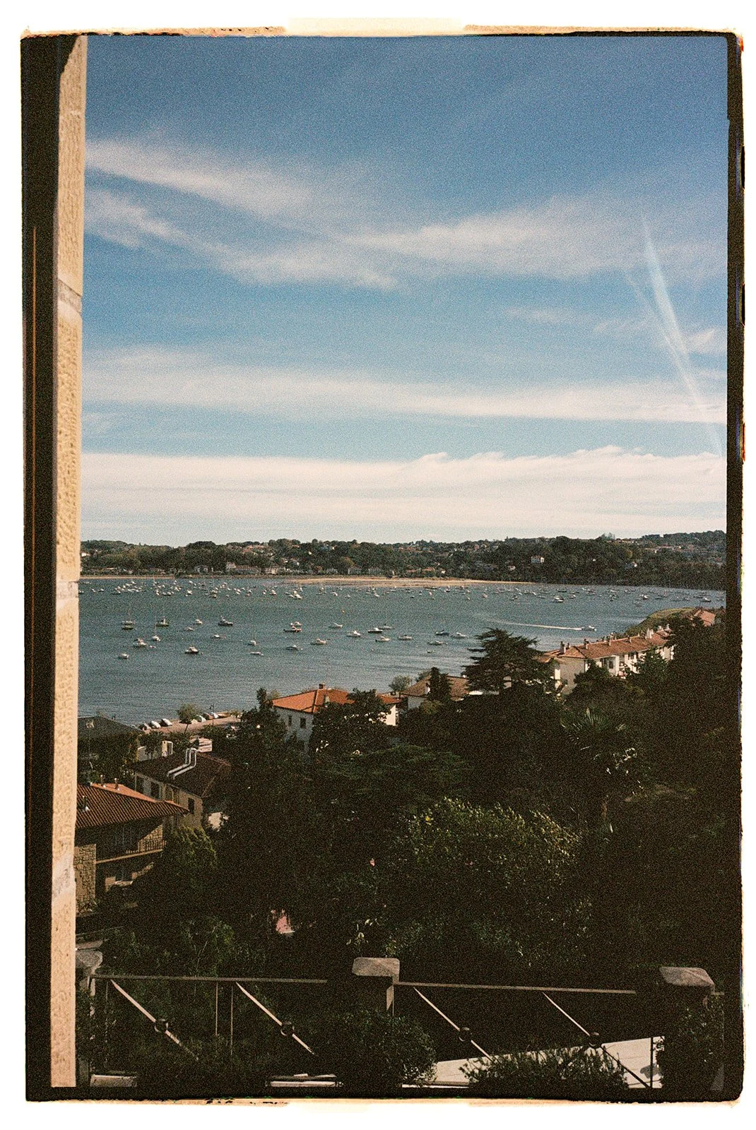 A scenic view of a river with boats, seen from a window with a stone frame, under a partly cloudy sky. Houses and trees are visible along the riverbank.