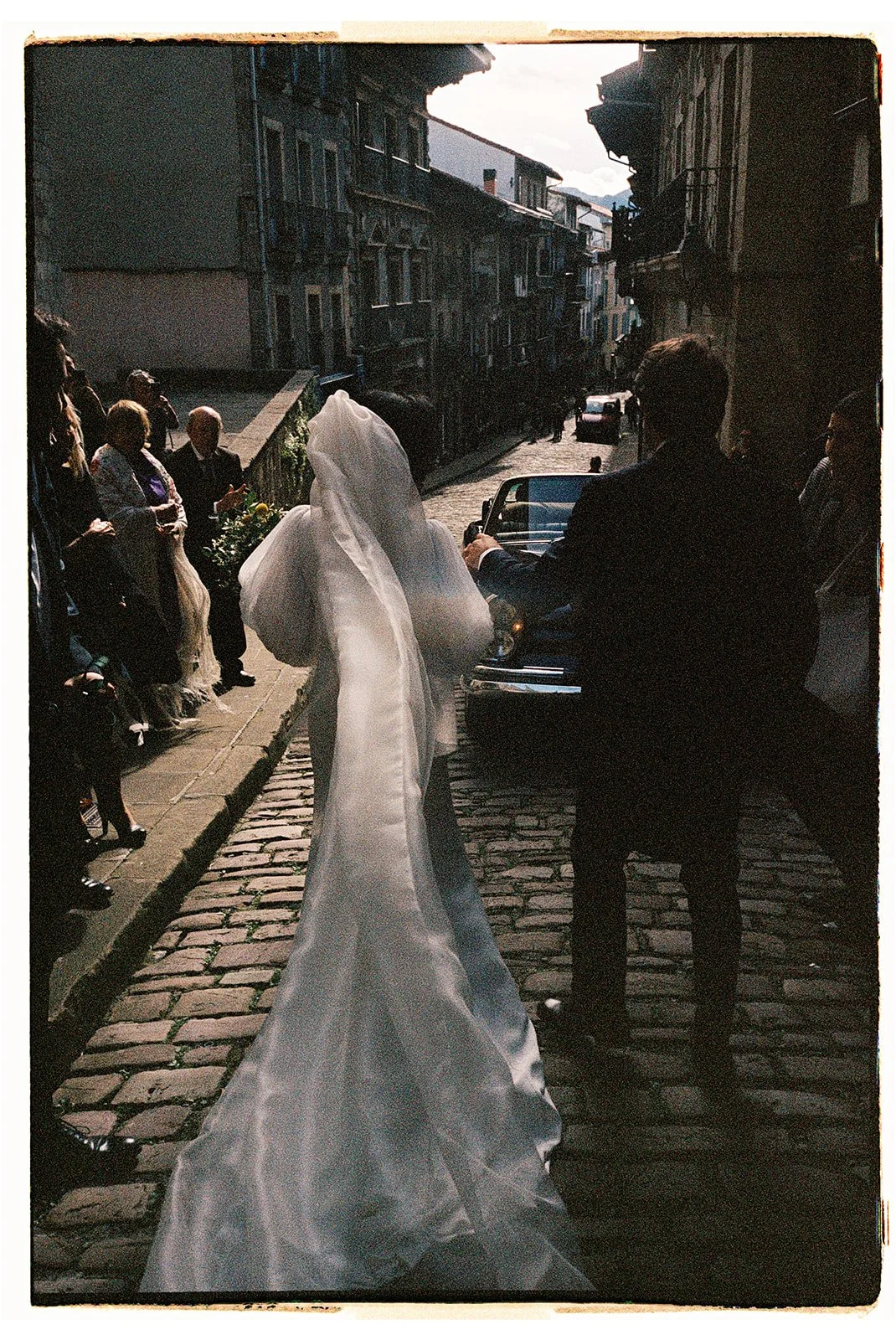 A bride in a wedding gown and veil walking on a cobblestone street toward a man in a suit, with onlookers on the sidewalk, in a historic European city.