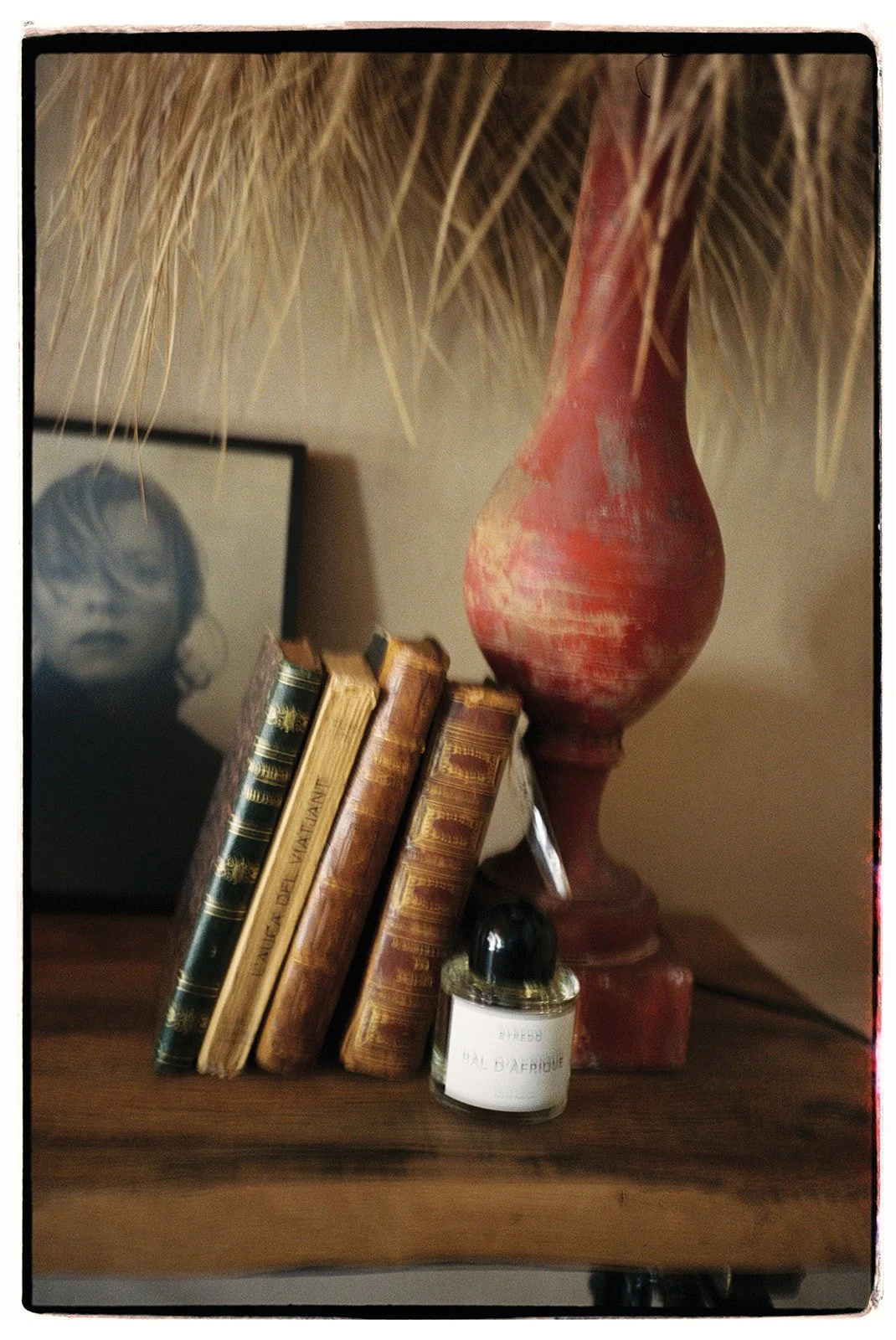 A wooden table with four old books, a small bottle of perfume, a large red vase, and a black-and-white framed photo of a person with sunglasses and light-colored hair. There are straw decorations hanging above.