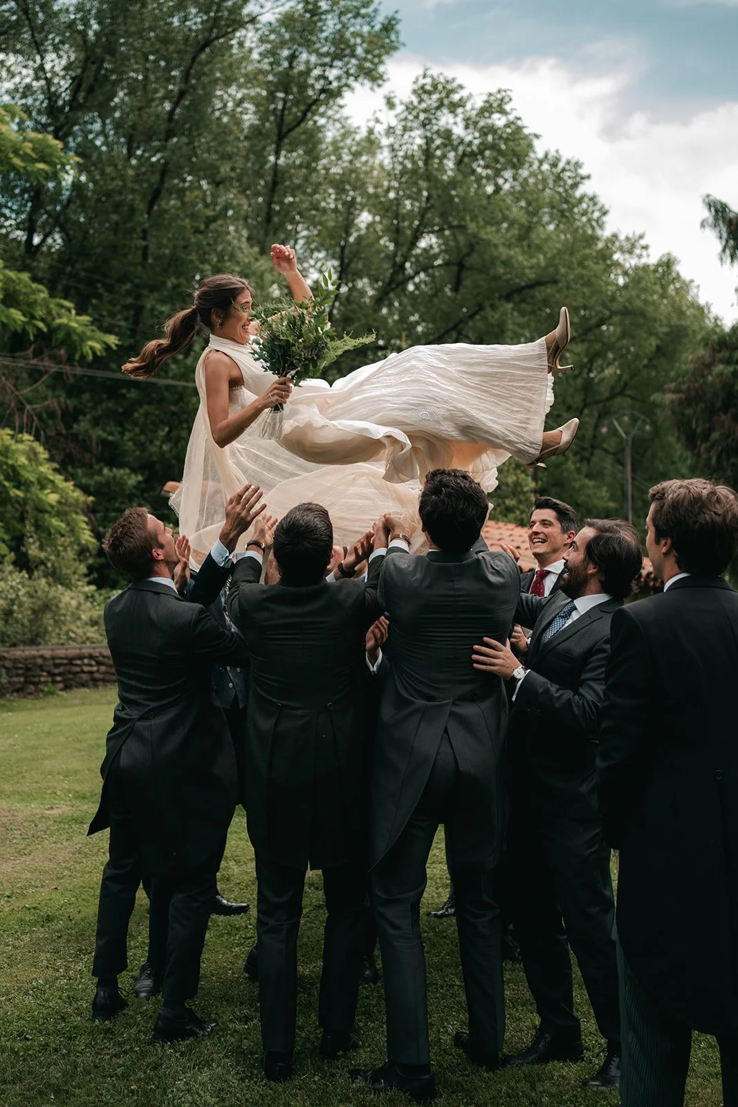 A bride in a white wedding dress is being lifted by a group of men in suits outdoors. She holds a bouquet of flowers and appears joyful as she is being thrown in the air among green trees and cloudy sky.