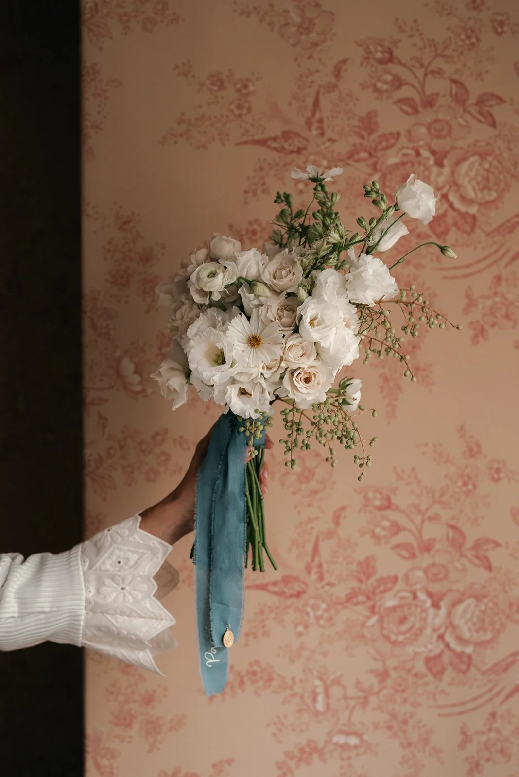 A hand holding a bouquet of white flowers with a blue ribbon against a pink floral patterned wallpaper background.