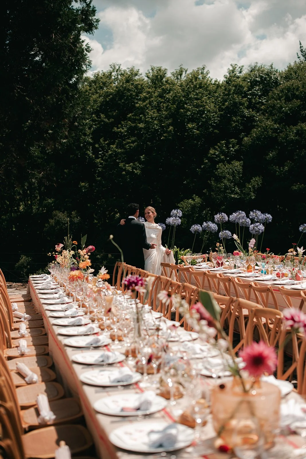 A bride and groom dancing at an outdoor wedding reception with a long, decorated table set with plates, glasses, and flowers, surrounded by lush green trees and a partly cloudy sky.