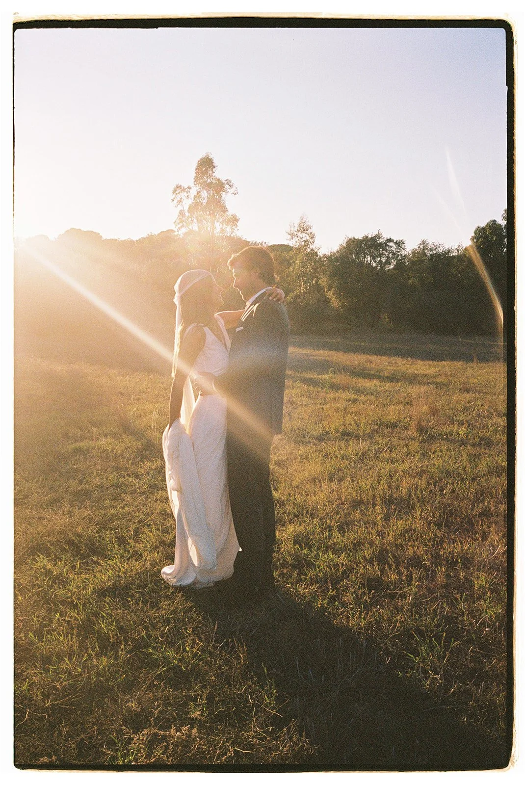 A couple is standing close together in a field during sunset, with the sun behind them creating a bright glow and lens flare.