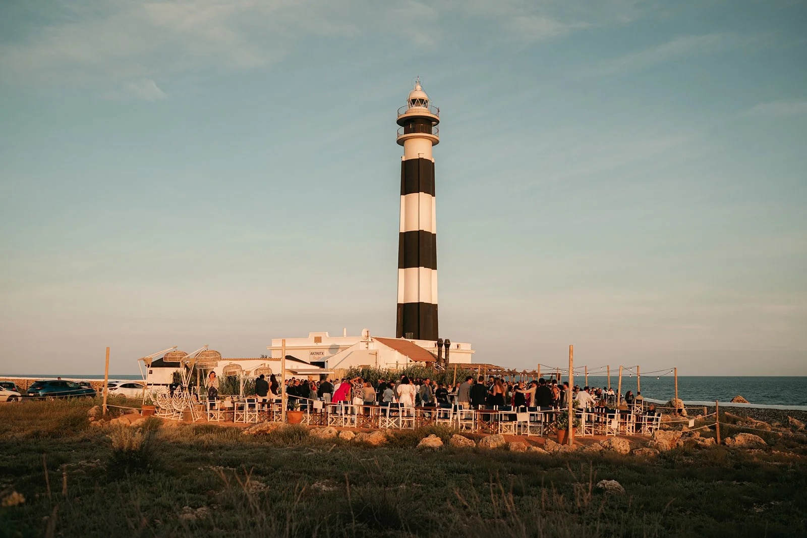 Lighthouse with black and white horizontal stripes near the ocean at sunset, with people gathering outside a building at its base
