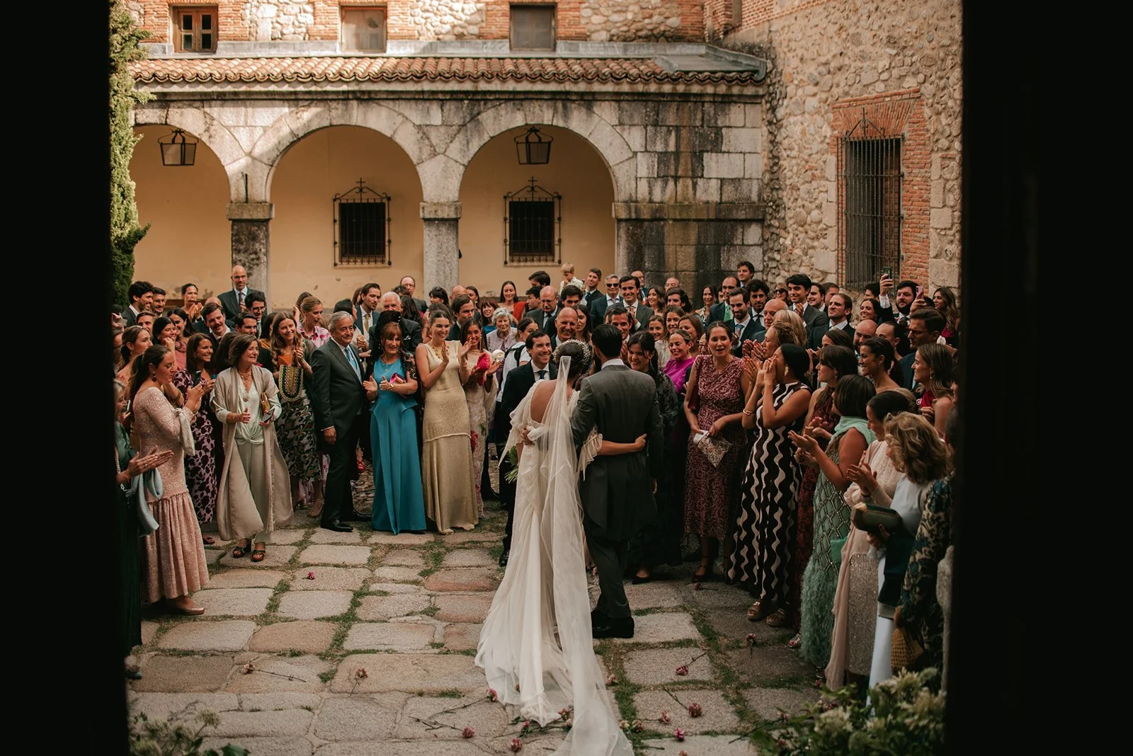 A wedding ceremony happening outdoors in a courtyard, with a bride and groom standing together, surrounded by many guests clapping and smiling.