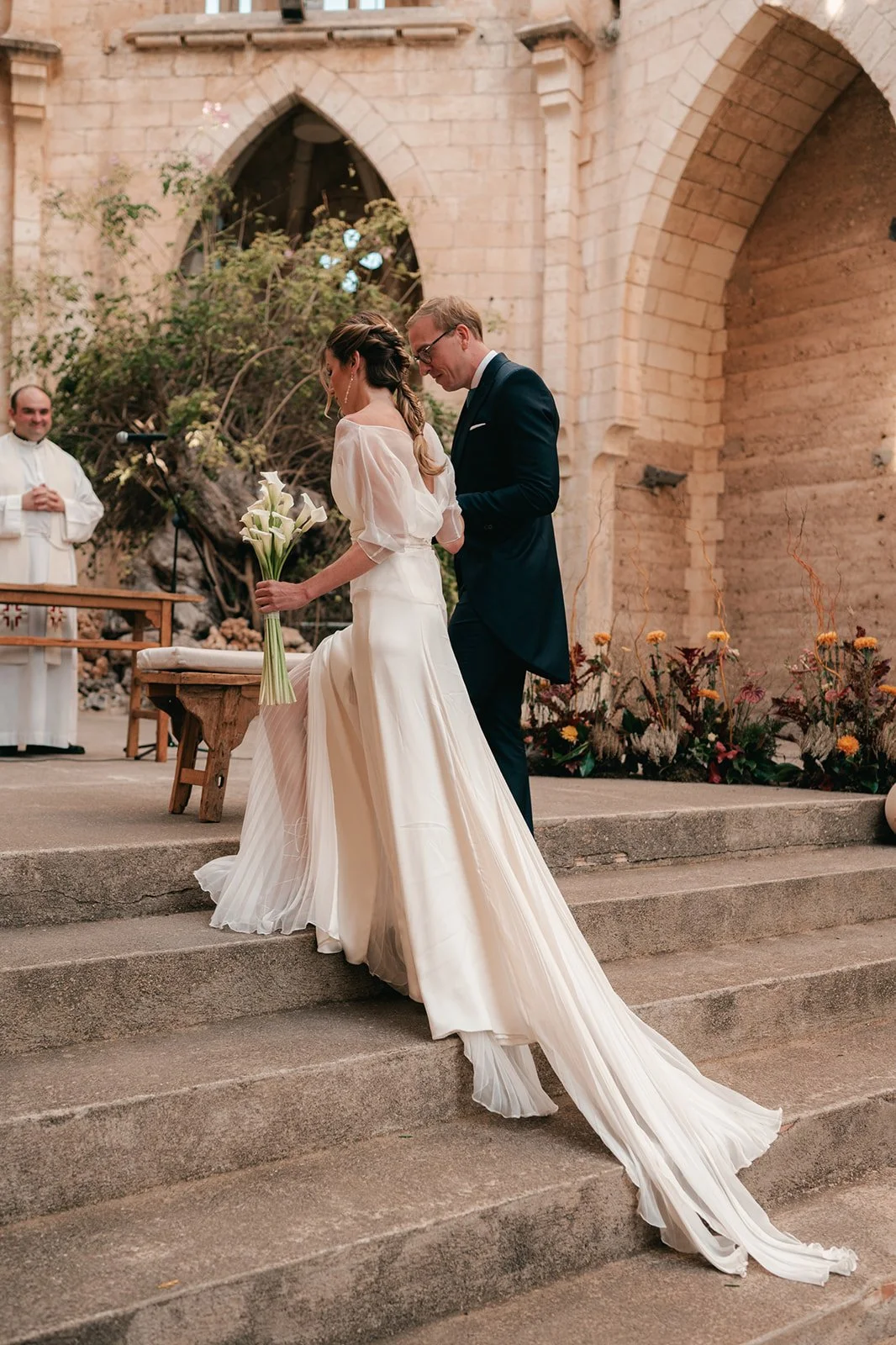 A couple getting married at an indoor church, with the bride holding a bouquet of white calla lilies, as the officiant looks on, flowers decorate the setting.