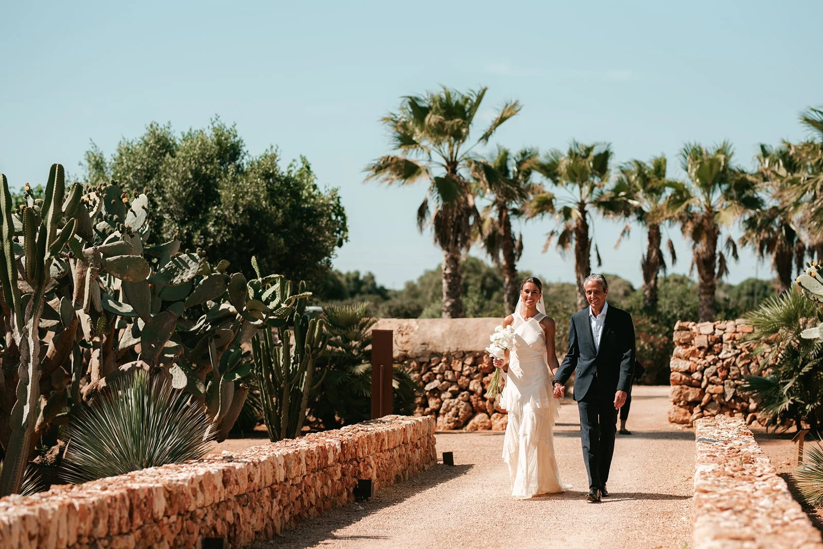 A bride in a white wedding dress holding a bouquet walks with a man in a black suit through a desert garden with cacti and palm trees under a clear blue sky.