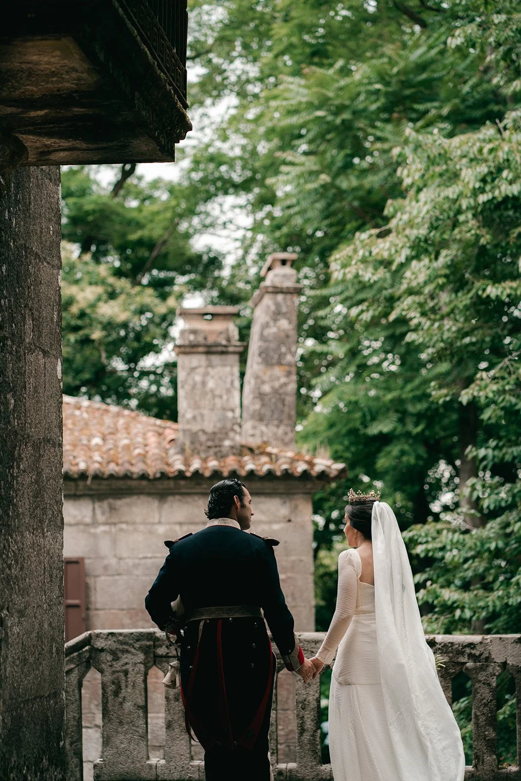 A bride and groom holding hands, standing outdoors on a stone balcony surrounded by lush green trees, with an old stone building with chimneys in the background.