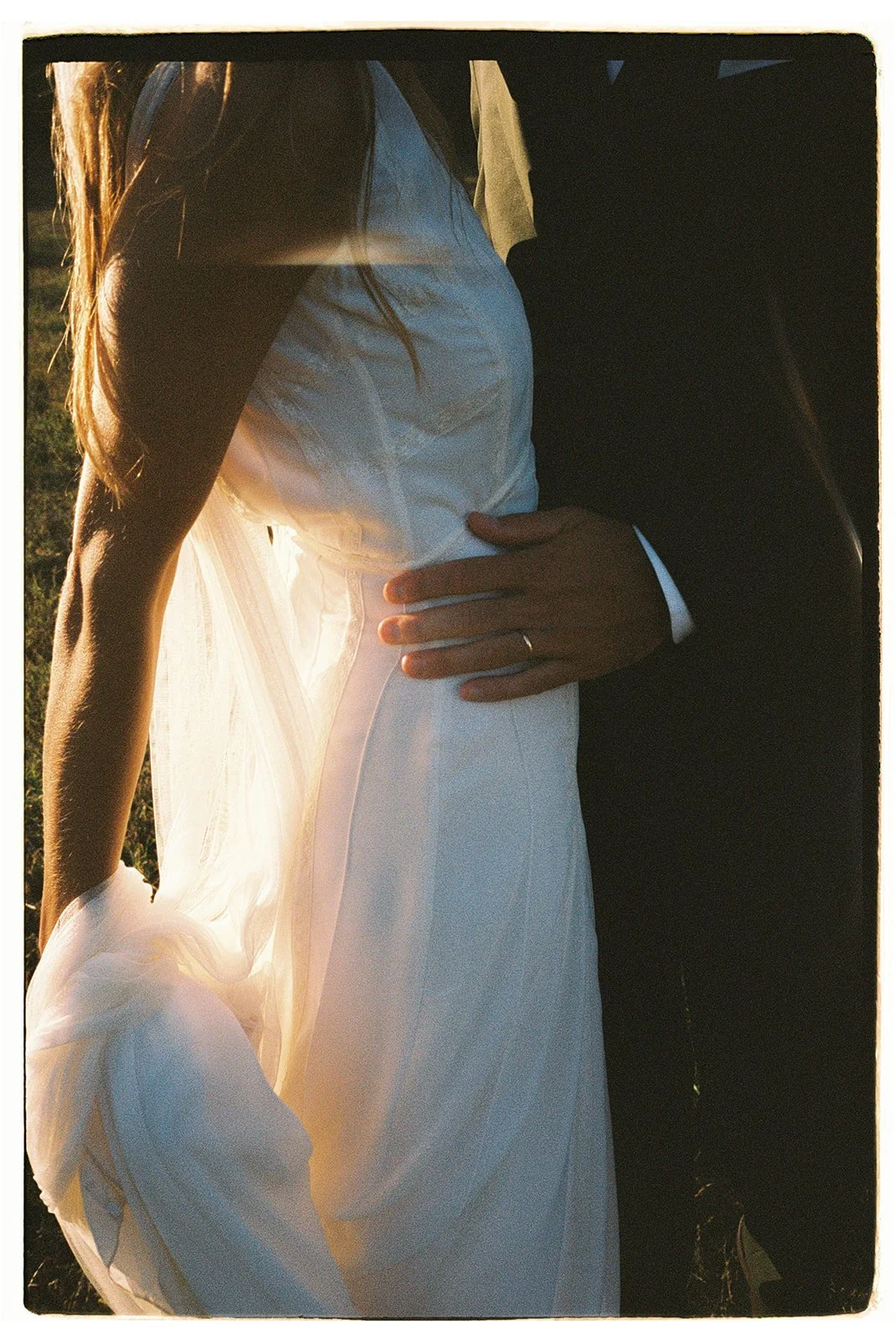 Close-up of a bride and groom holding each other outdoors during sunset, with sunlight shining through the bride's white wedding dress.