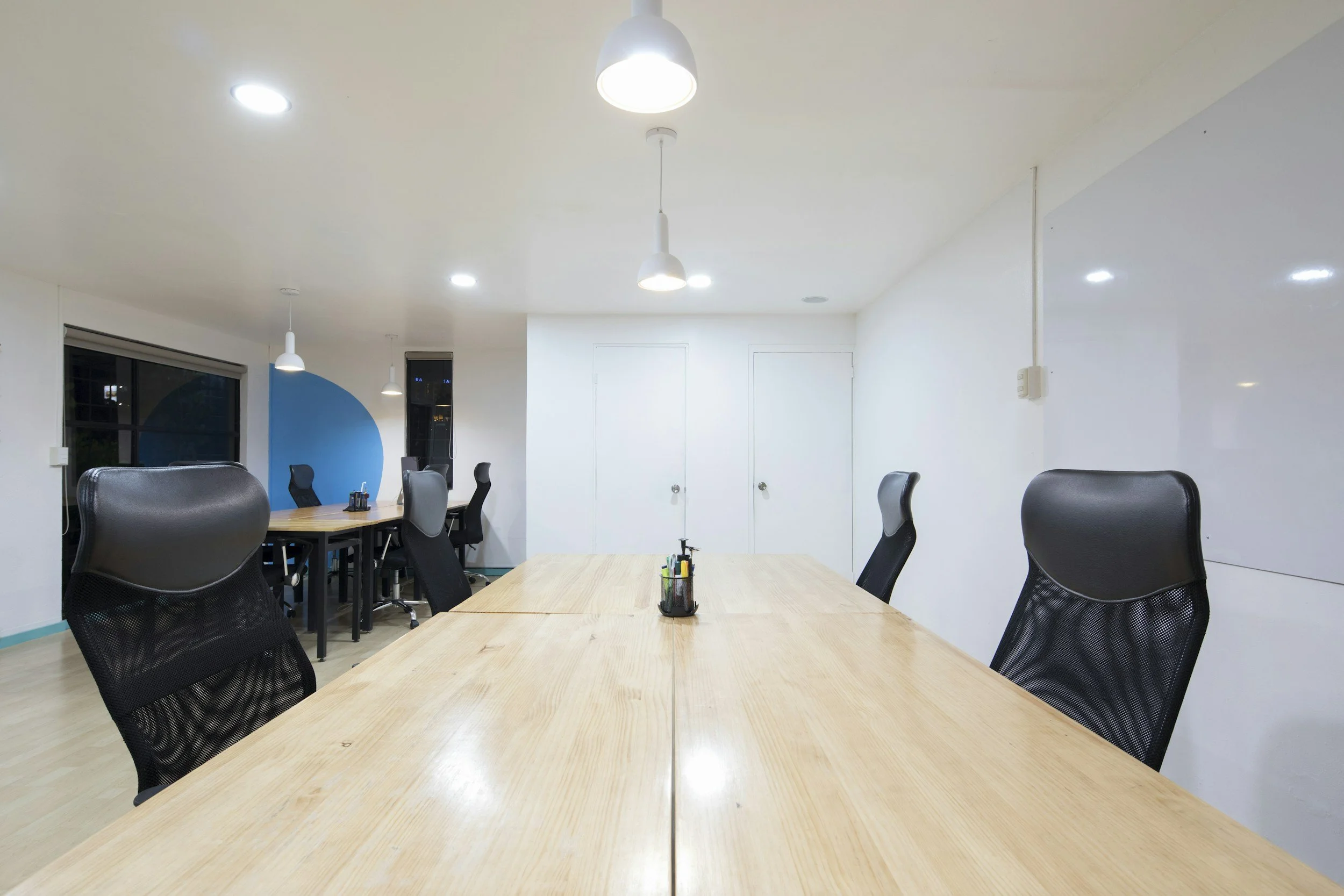Empty conference room with wooden tables, black ergonomic chairs, white walls, light fixture, whiteboard, and window with city view.