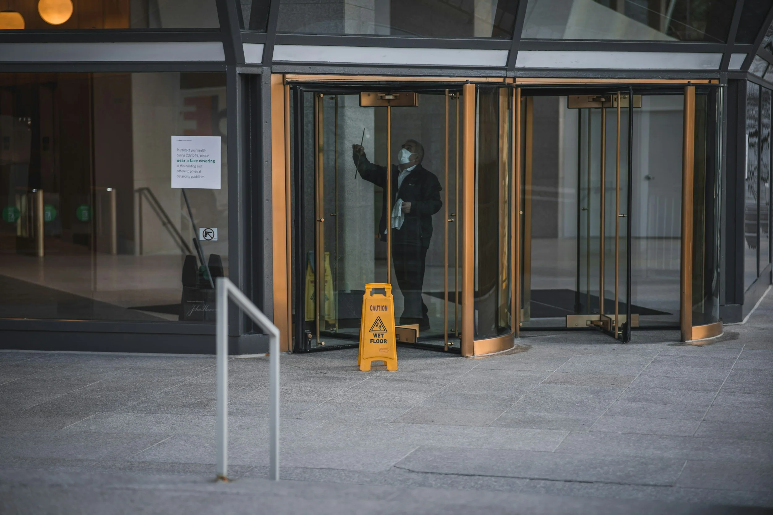 Man wearing a face mask cleaning glass revolving door at building entrance with warning sign for wet floor.