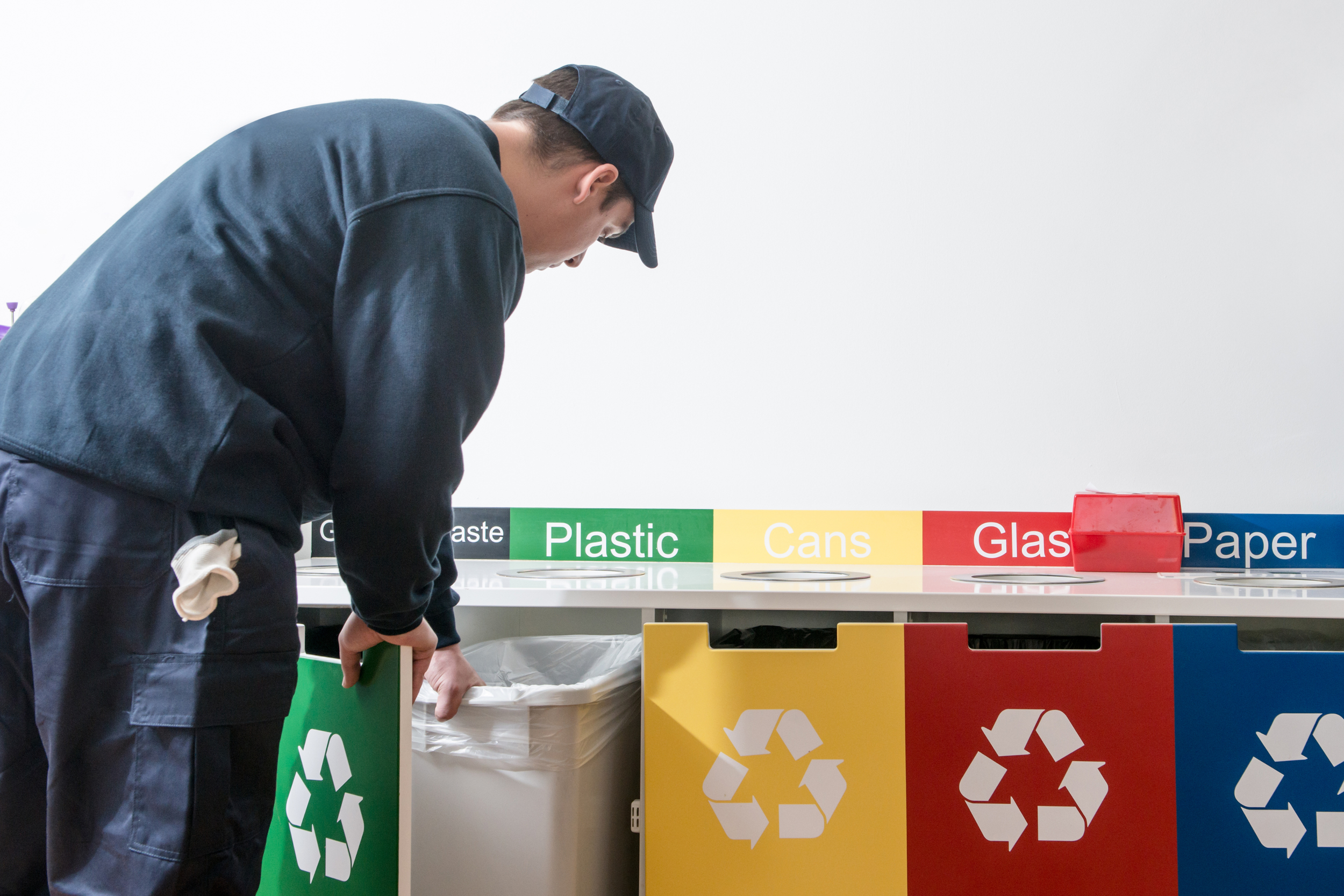 Person throwing recyclables into recycling bins labeled for plastic, cans, glass, and paper in a waste sorting area.