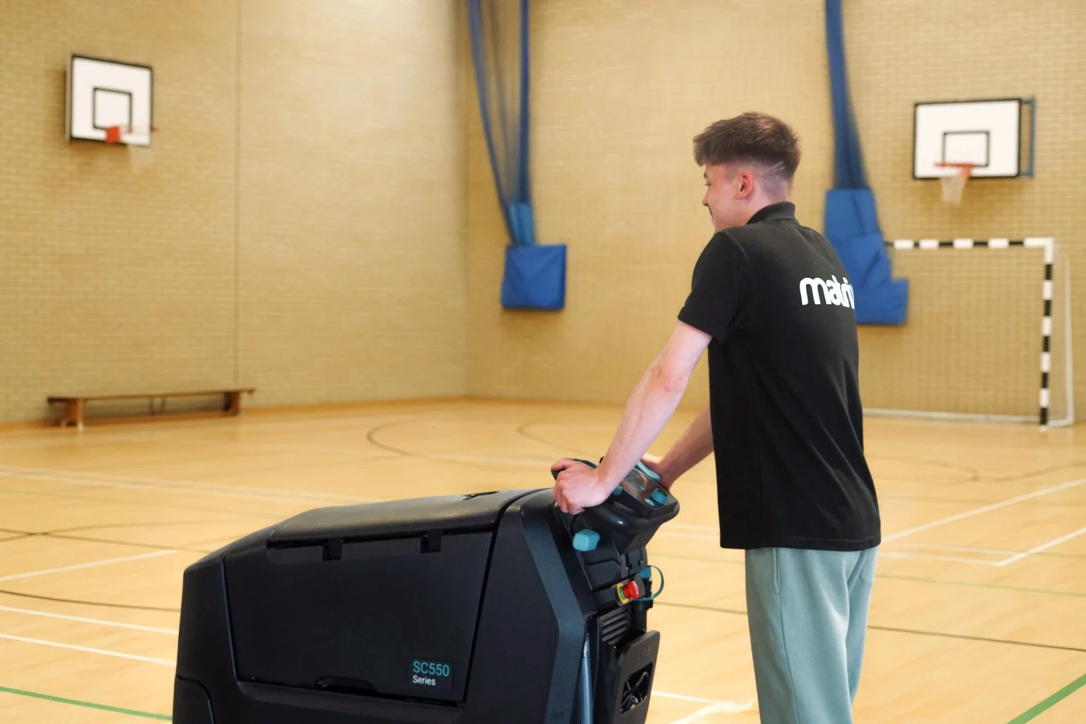 A young man in a black shirt pushing a large floor cleaning or sanitizing machine in a gymnasium with wooden floors, basketball hoops, and a goalpost in the background.