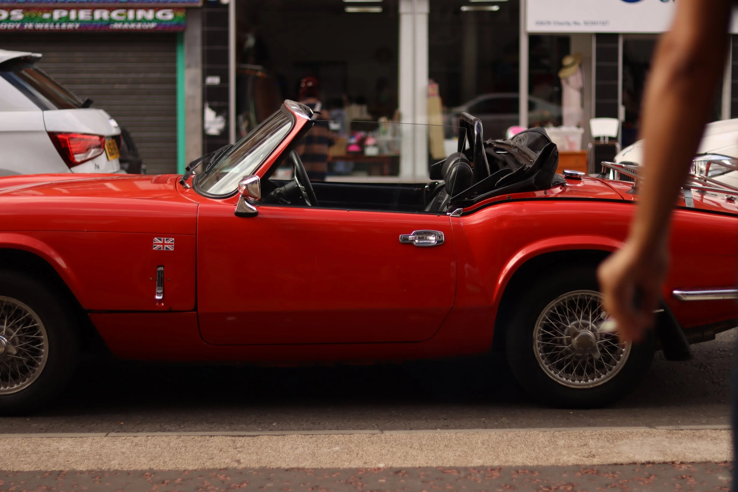 Red vintage convertible sports car parked on city street with a sticker of the Union Jack flag on the side, people and shops in the background.