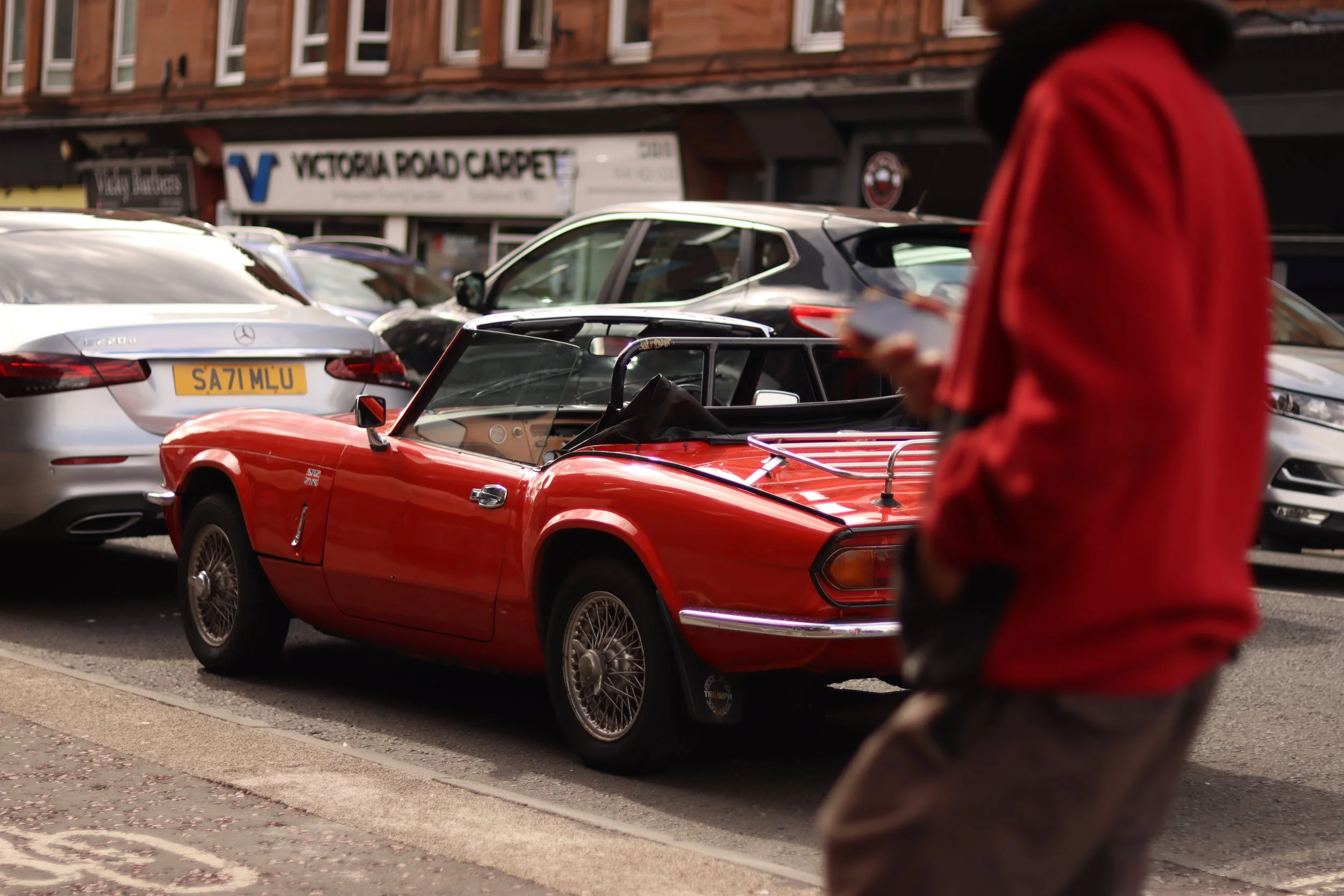 A vintage red Triumph convertible parked on a city street, with modern cars and a pedestrian in red jacket nearby.