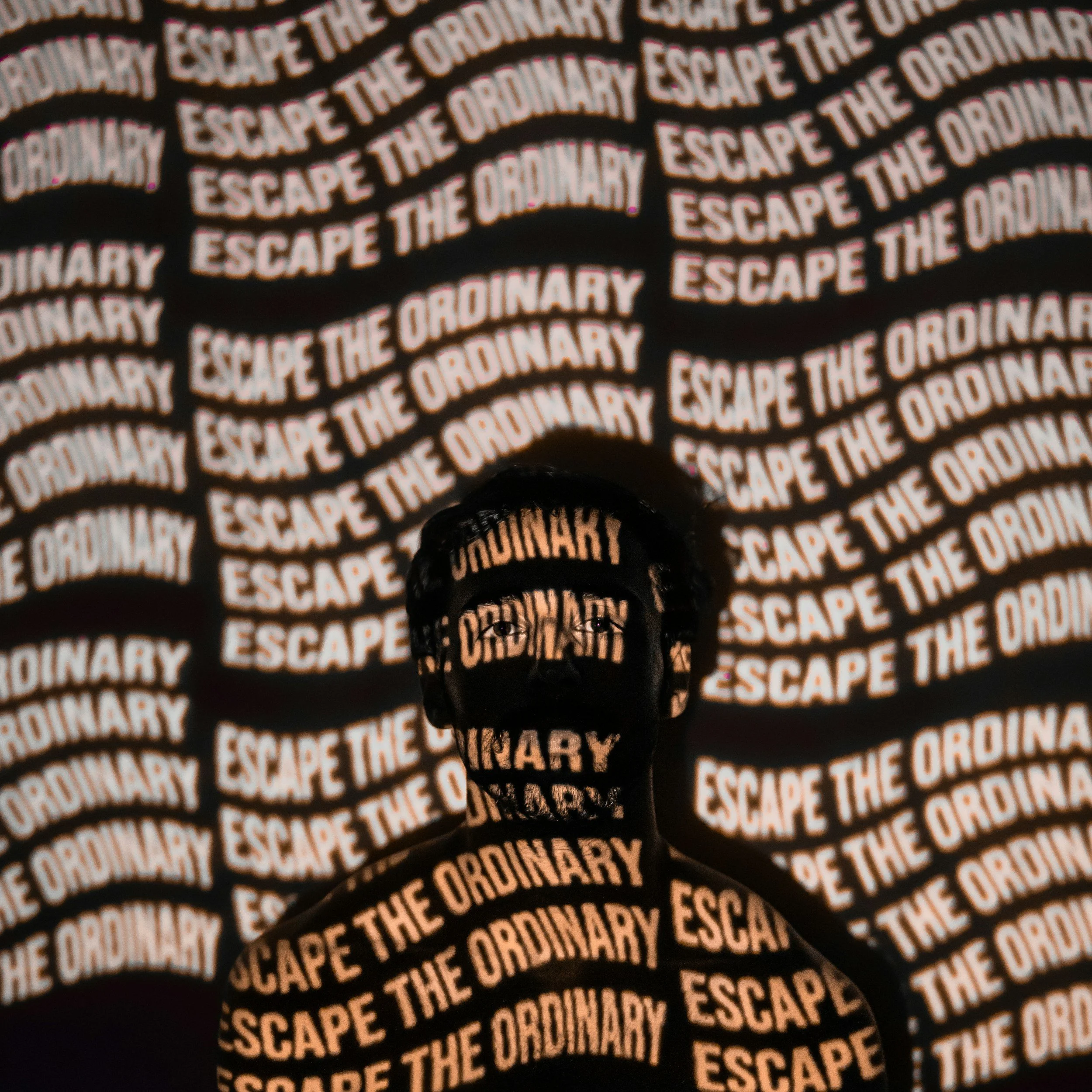 A person with a lit head and face, with the phrase "ESCAPE THE ORDINARY" projected onto their face and body, and the background filled with the same phrase repeated numerous times.