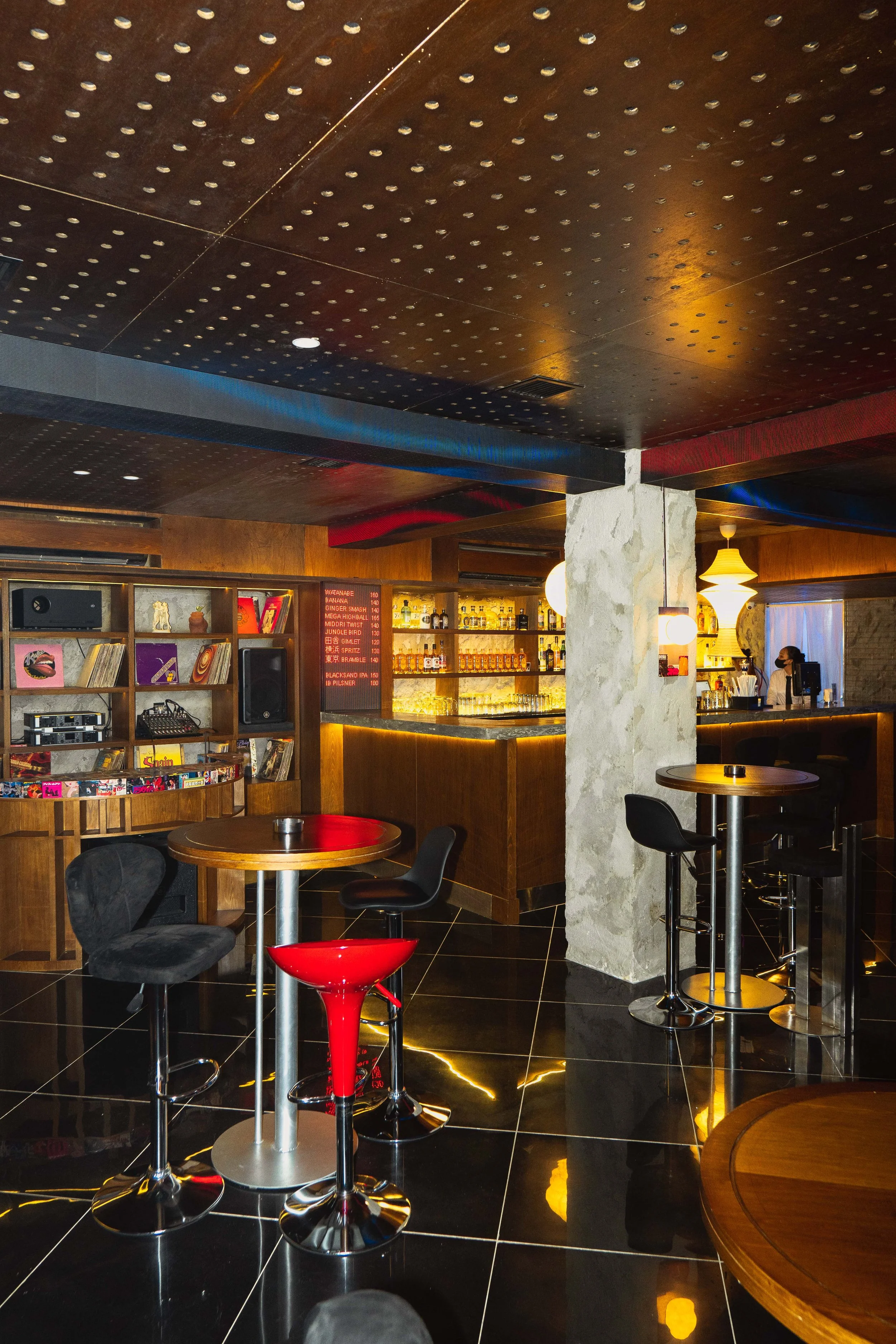 Interior of a dimly lit bar with a wooden counter, shelves of liquor bottles, and a music record shelf. There are round tables with chairs and bar stools, with warm lighting and a reflective black tile floor.