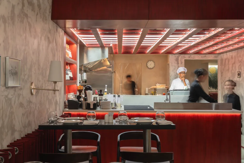 Interior view of a restaurant kitchen seen through an open window, with staff preparing food and a chef wearing a white uniform and hat.