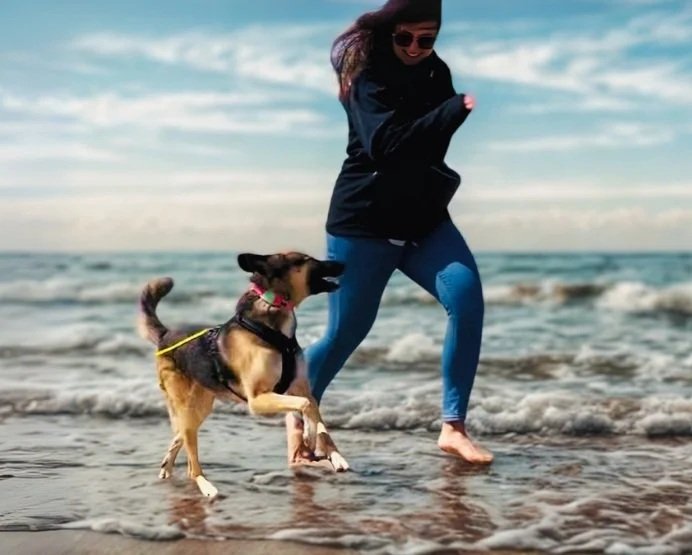 Eine Frau und ein Hund laufen barfuß am Strand entlang, das Meer und der Himmel im Hintergrund.